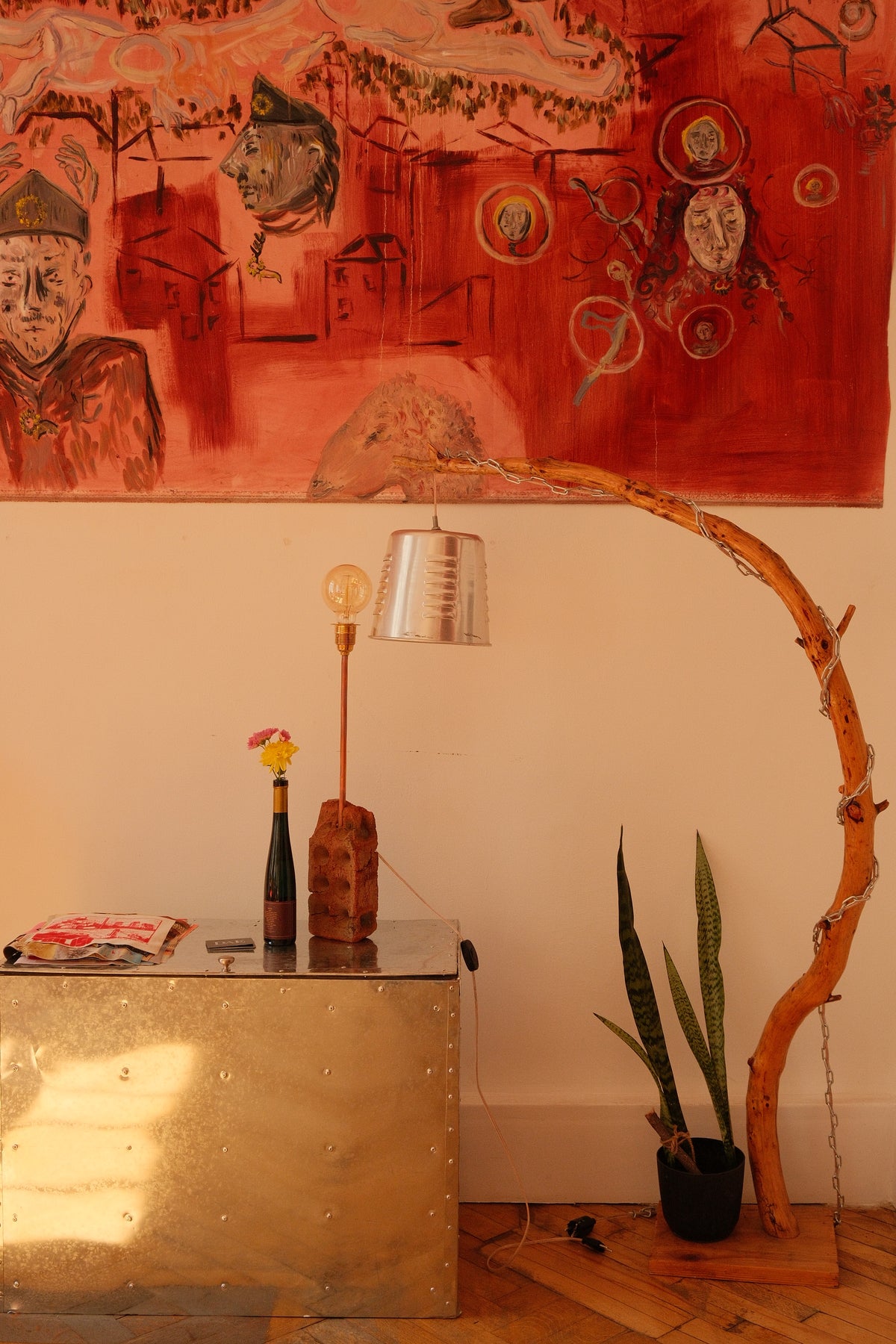 A styled interior corner featuring a large red abstract painting on the wall, a metallic trunk table, a curved wooden floor lamp, a small table lamp, a potted snake plant, and a bottle with a flower.