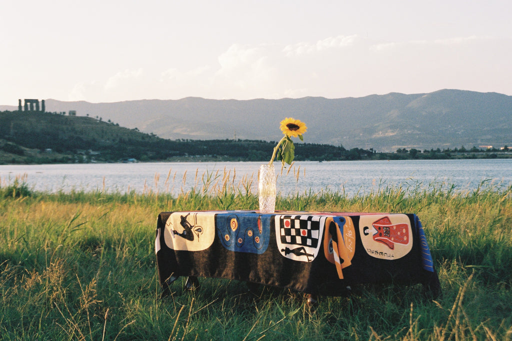 Tablecloth “Table of Figures” by Nino Bakuradze – sublimation-printed fabric with vibrant illustrated characters. Tablecloth in nature, near lake