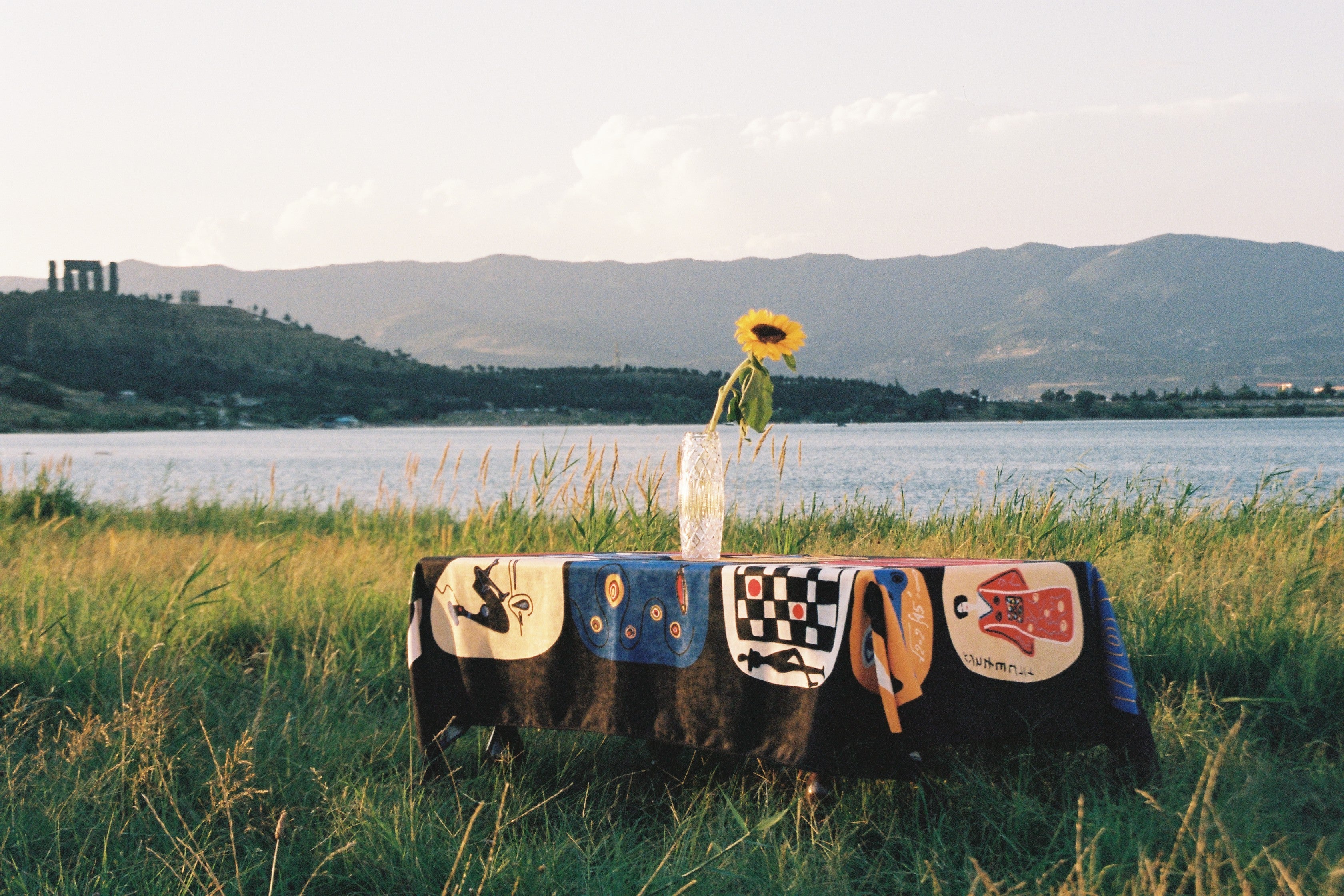 Tablecloth “Table of Figures” by Nino Bakuradze – sublimation-printed fabric with vibrant illustrated characters. Tablecloth in nature, near lake