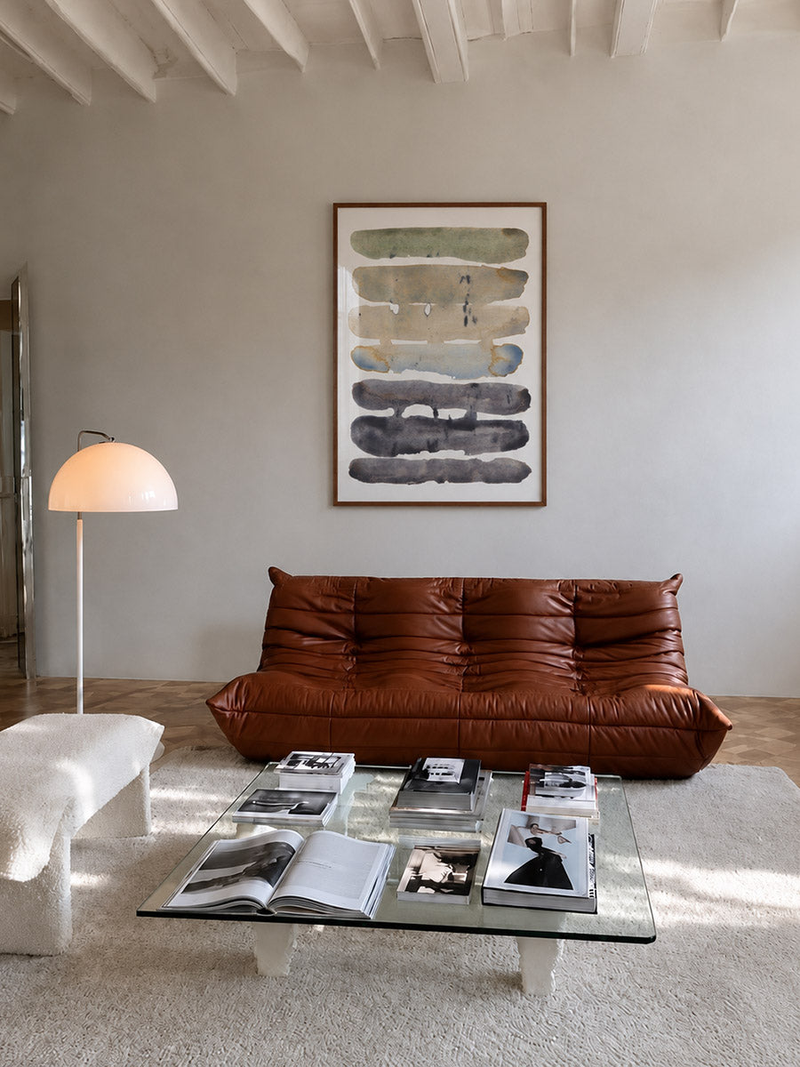 A modern living room scene featuring a brown leather sofa under a framed abstract poster by DAE on a light gray wall. A glass coffee table with magazines sits on a white rug, and a white floor lamp is on the left.