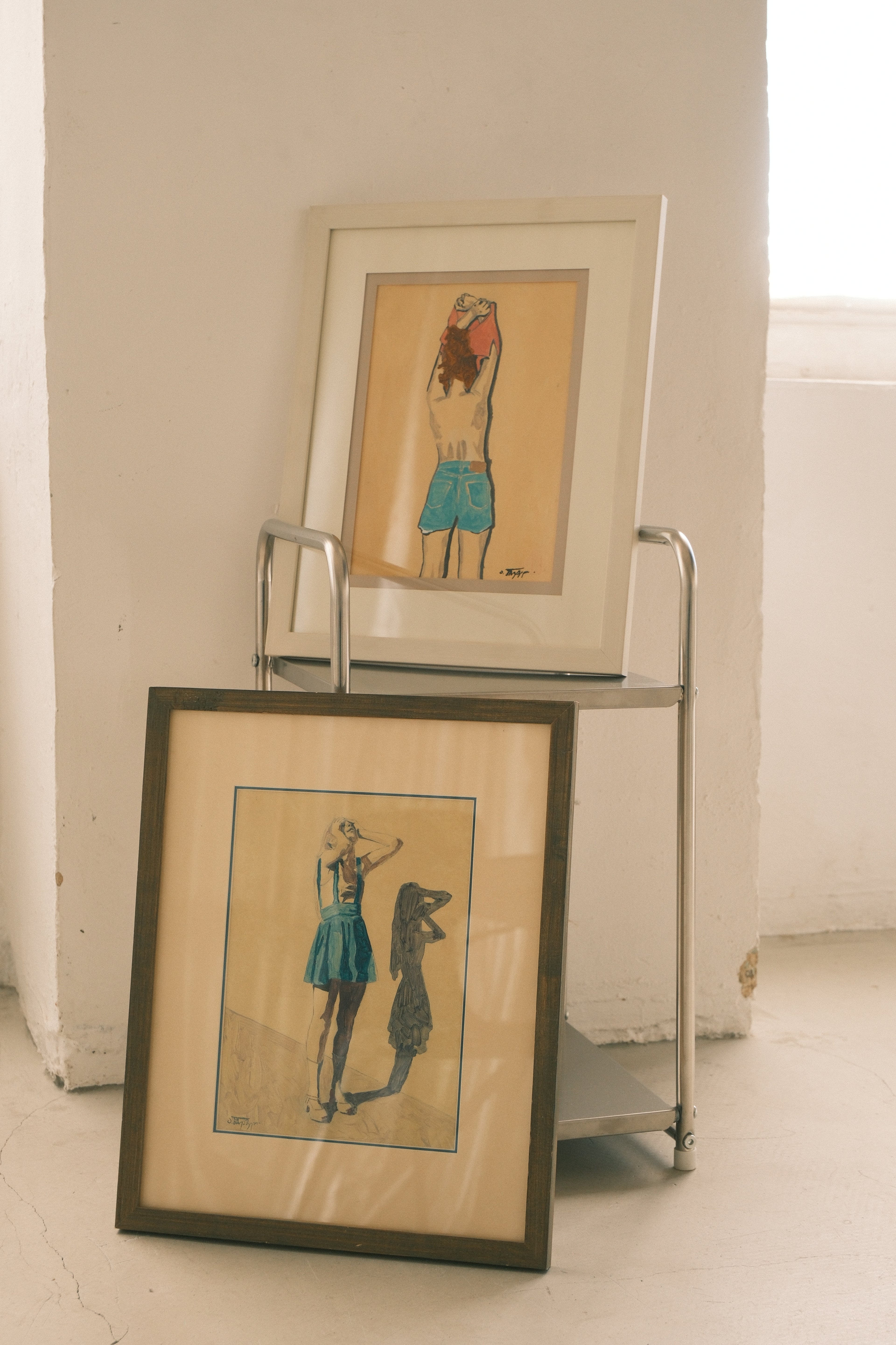 Two framed artworks on a metal cart against a white wall.