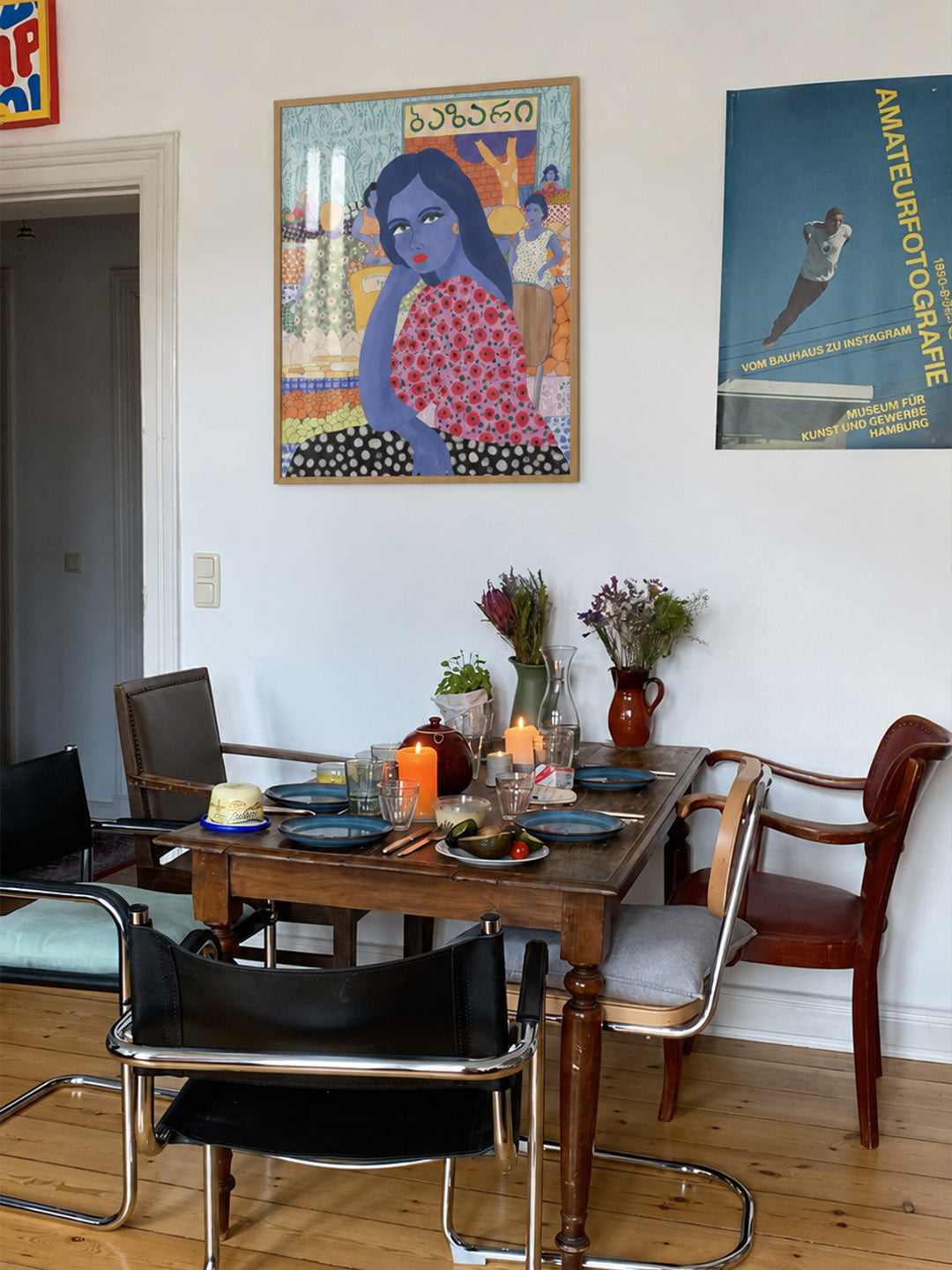 A dining room scene with a wooden table set for a meal, surrounded by several chairs. On the white wall behind are two framed posters: one colorful portrait-style print and one blue vintage-style poster with text.