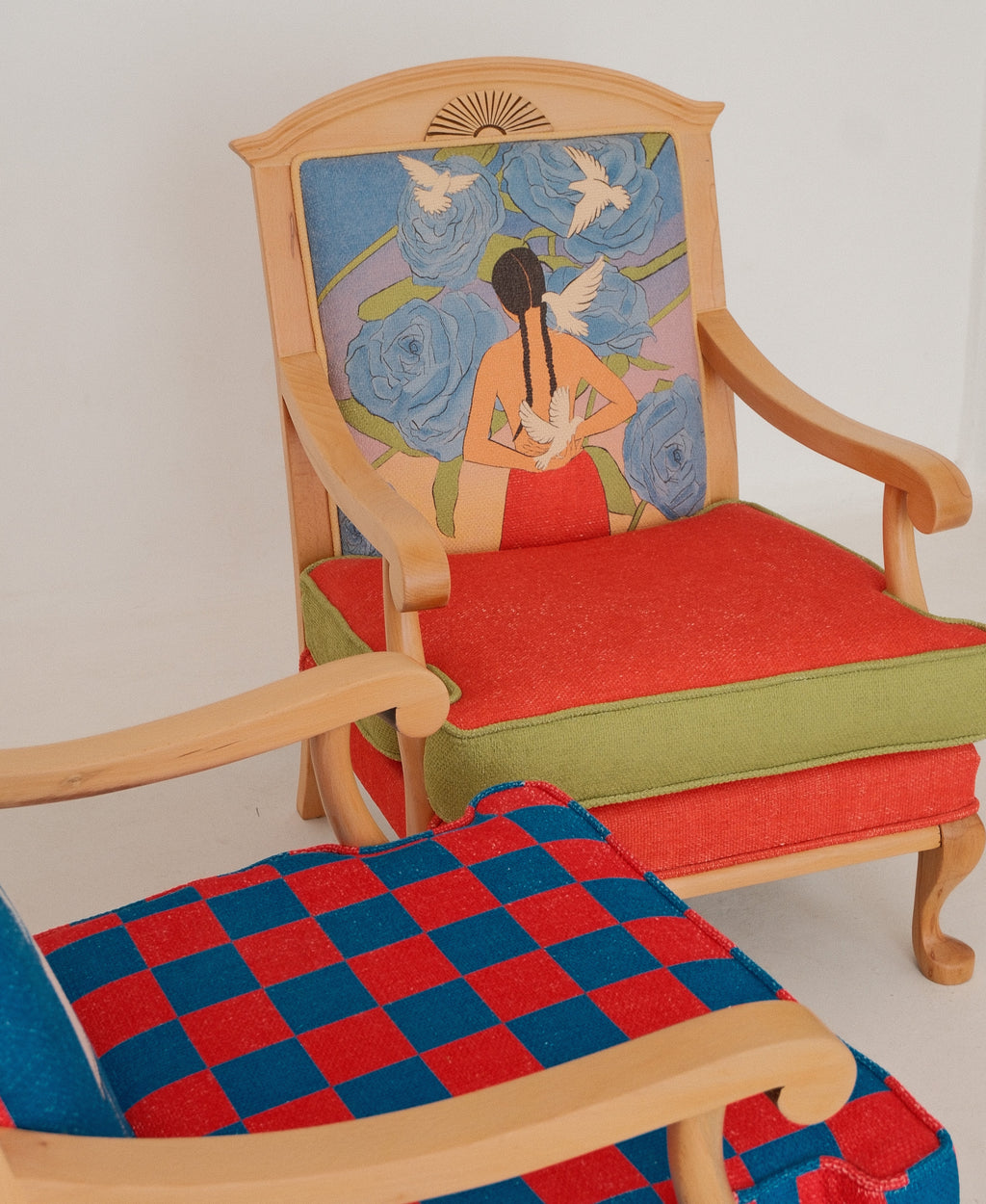 A wooden armchair with a colorful floral and bird upholstered backrest and red seat cushions, shown alongside another chair with a red and blue checkered cushion in the foreground.