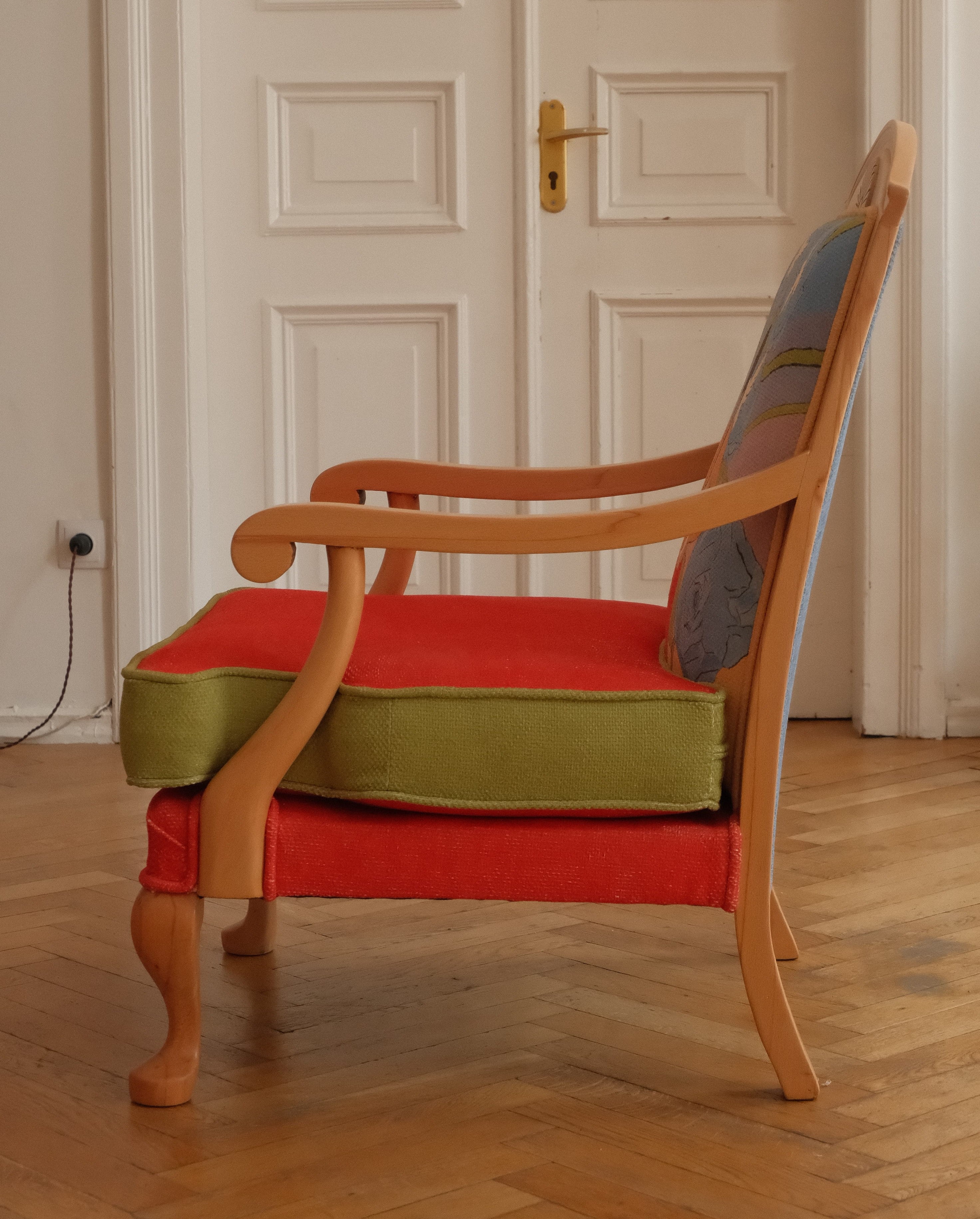 A wooden armchair with a red upholstered seat and back cushion, olive green side bolsters, and a blue-gray patterned backrest, shown in a room with a wood floor and paneled doors.