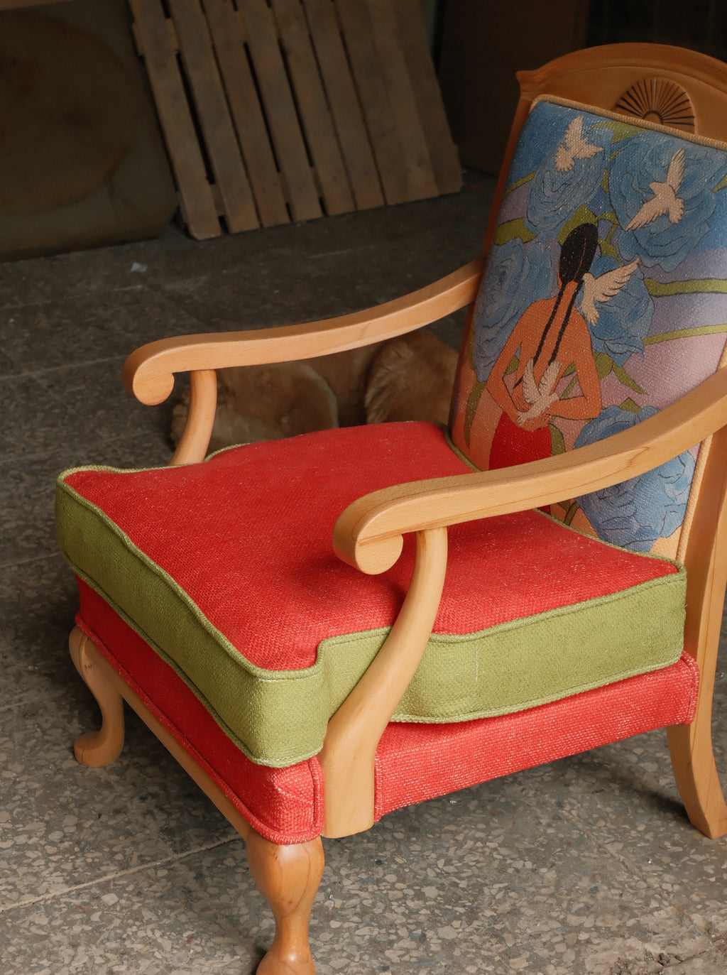 A wooden armchair with a red upholstered seat cushion and green side bolster, featuring a blue floral backrest with a stylized figure illustration.