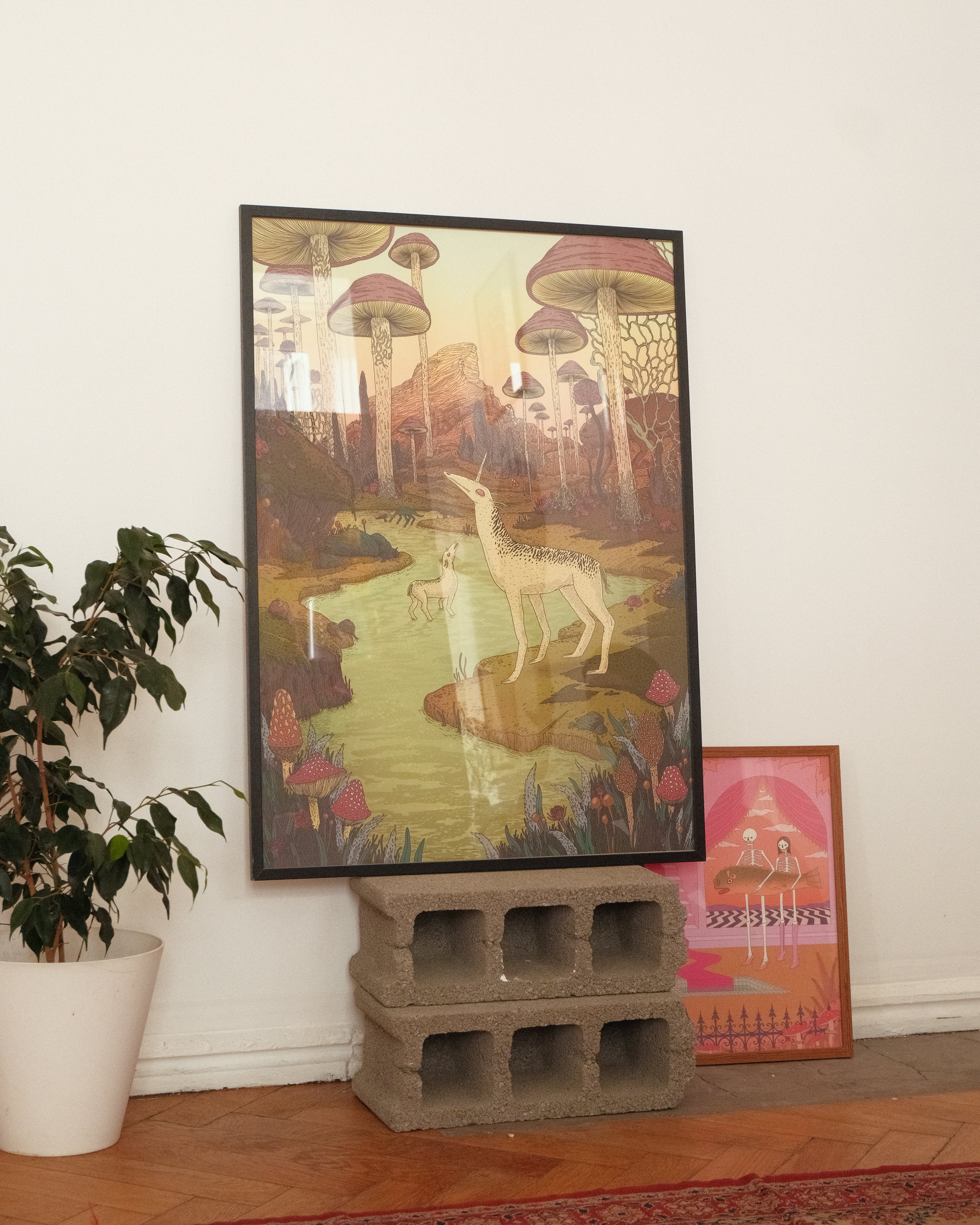 Framed artwork of a deer in a fantasy landscape on a white wall with a plant and another piece of art in the background.