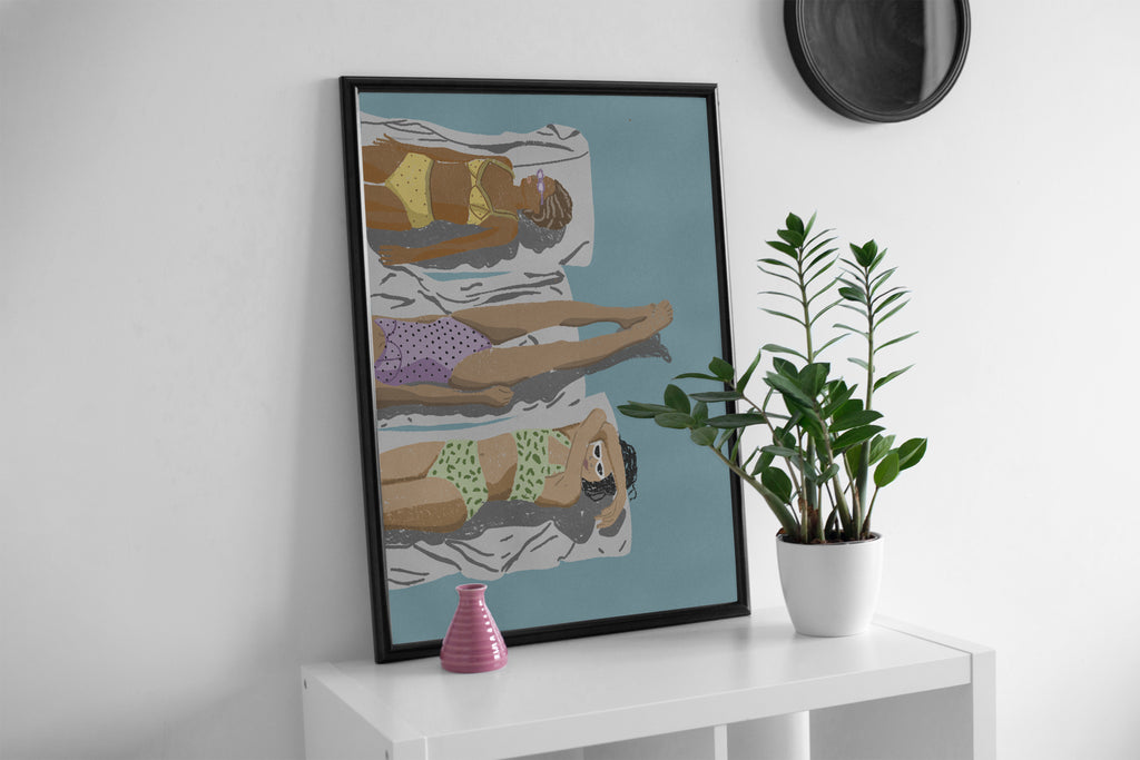 A framed poster of three stylized figures relaxing on beach chairs against a blue sky and turquoise sea background, hung on a white wall.