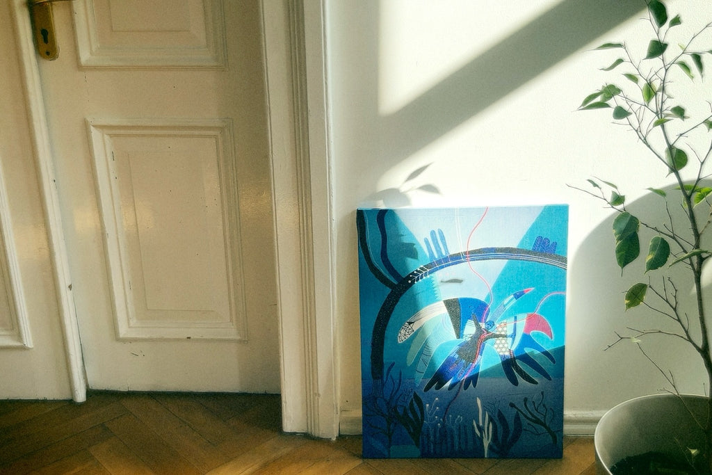 A canvas print with an abstract blue design, hanging on a white door in a room with wooden flooring. There is a plant to the right of the canvas.