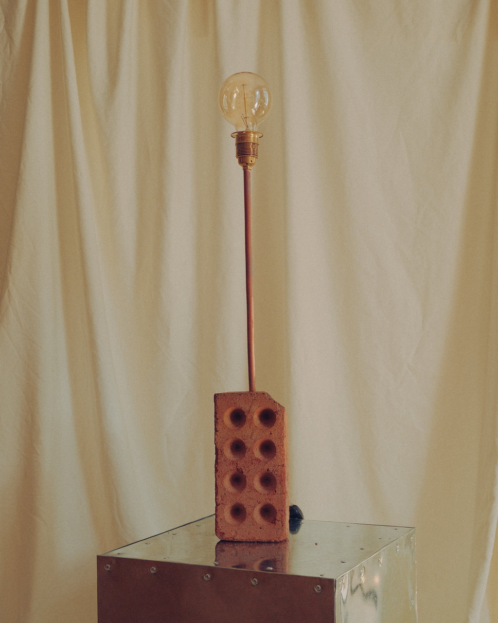 A unique table lamp featuring a brick base with a long copper-colored stem supporting a clear glass bulb, set on a reflective metal pedestal against a draped cream fabric backdrop.