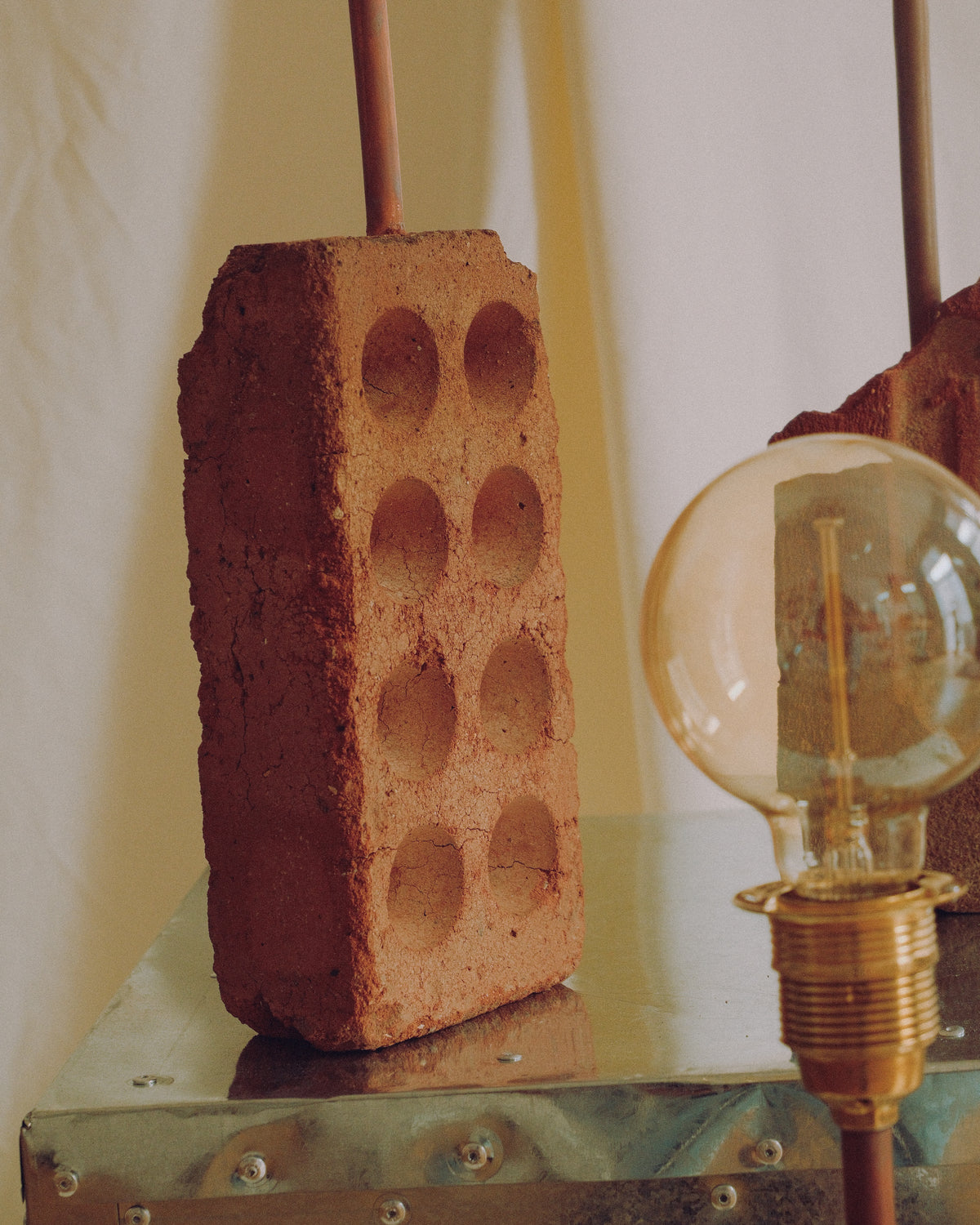 A rustic terracotta clay lamp holder shaped like a brick with circular indentations, holding a copper rod, styled next to an Edison bulb on a metal surface against a cream backdrop. handmade lamp by DAE