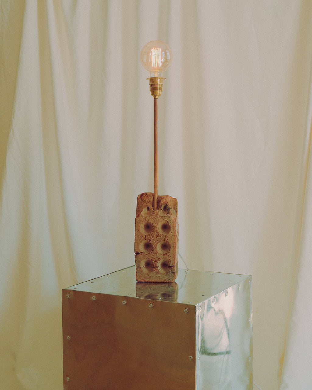 A rustic industrial-style table lamp featuring a weathered brick base with a central hole, a brass-finished metal stem, and a glowing clear glass bulb, displayed on a reflective metallic cube against a cream fabric backdrop.