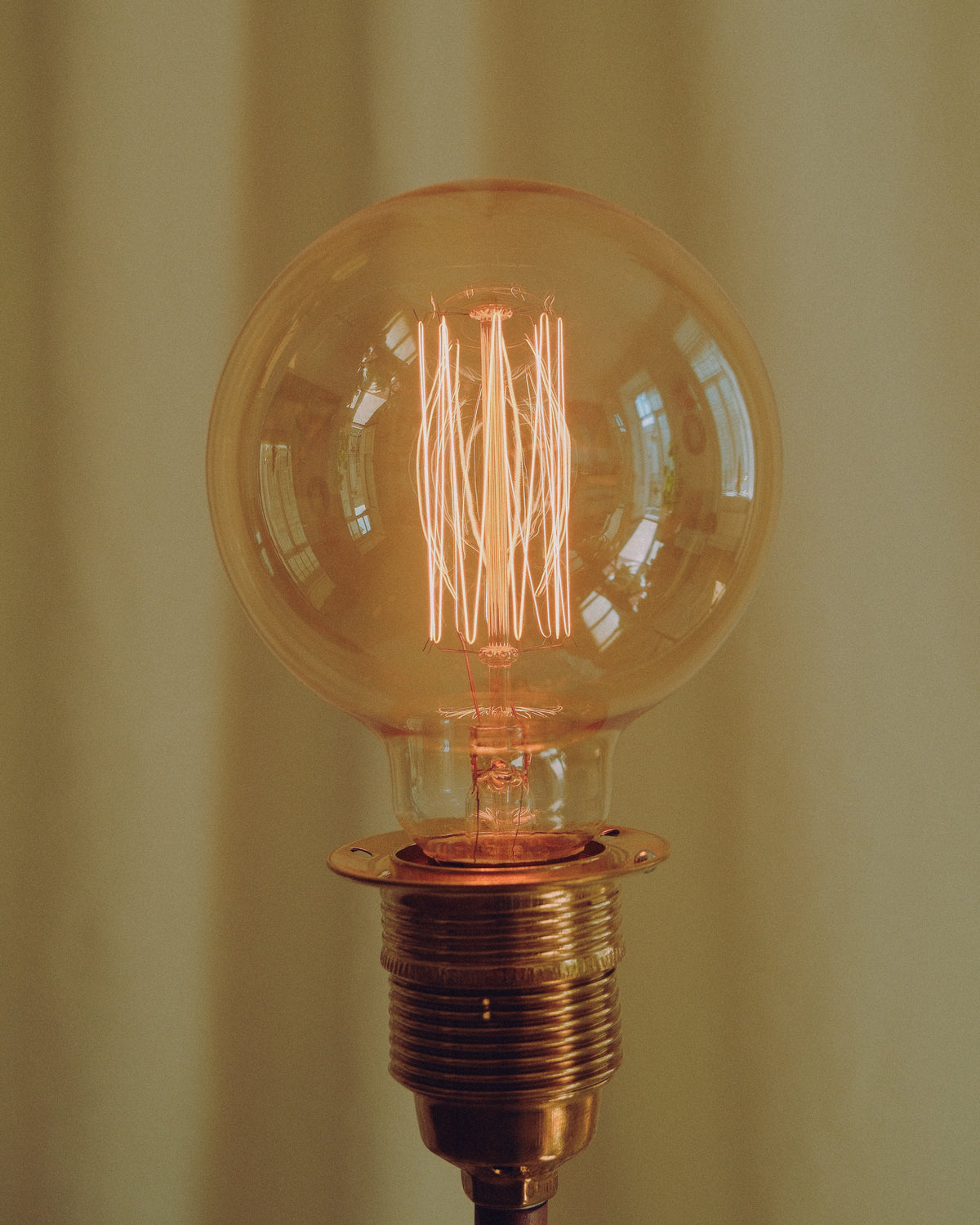 A large clear glass globe light bulb with a warm orange-tinted filament, mounted on a brass screw base against a softly lit beige background.