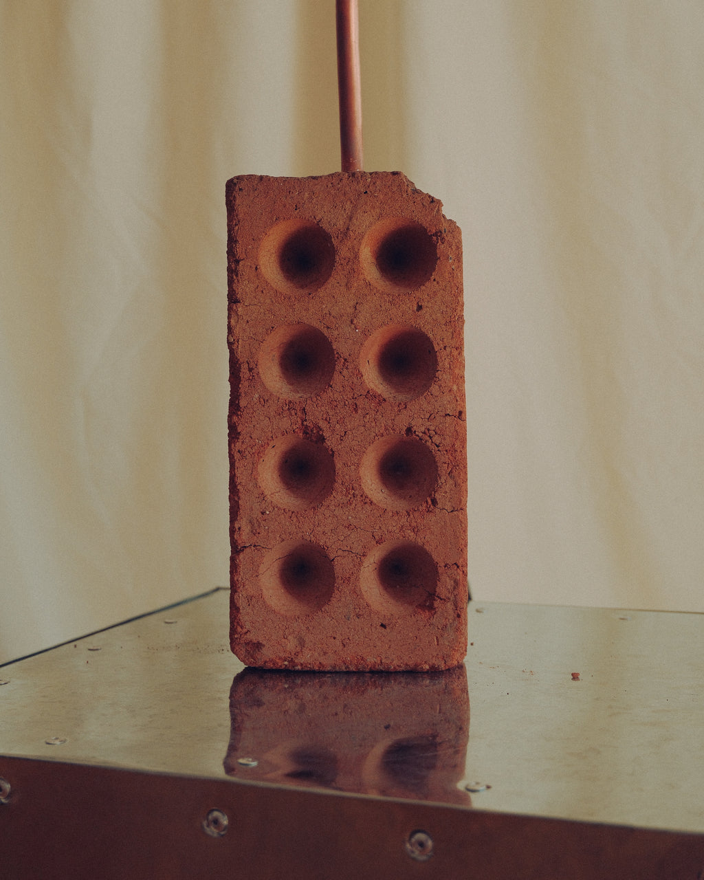 A single red clay brick with six circular holes, standing upright on a reflective metallic surface against a draped beige fabric backdrop.