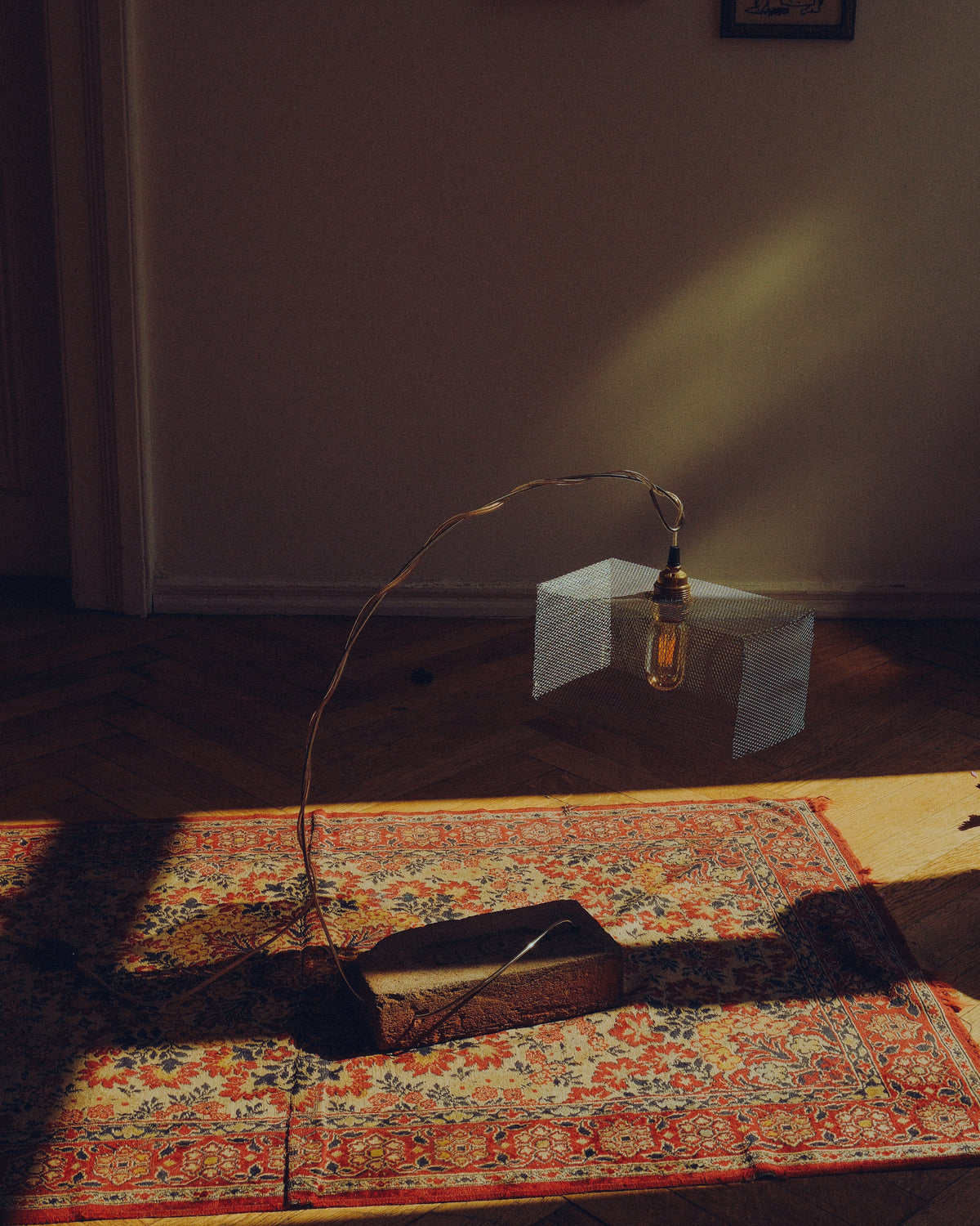 A rustic, industrial-style pendant light with a textured white rectangular shade, a long thin metal wire frame, and a visible Edison bulb, suspended over a patterned oriental rug in a dimly lit room.