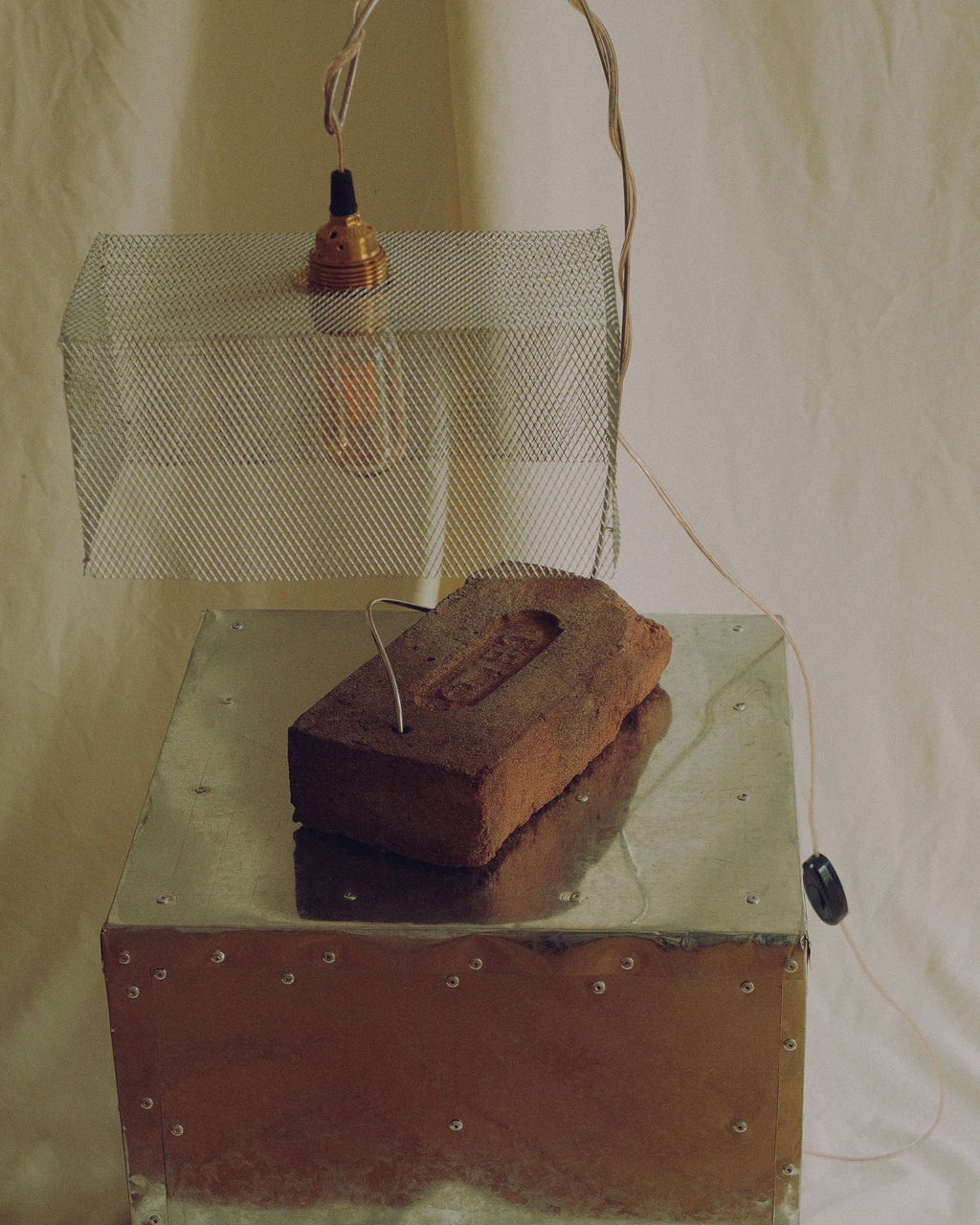 A rustic industrial-style pendant light with a wire mesh shade and exposed bulb, hanging above a metal box with a brick resting on it, photographed against a draped fabric backdrop.