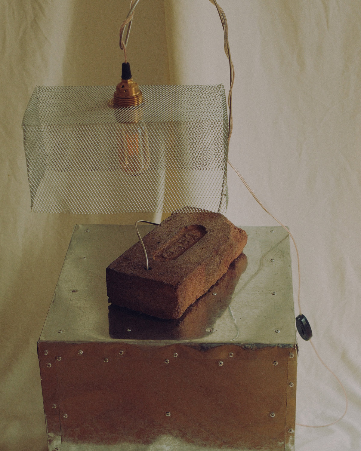 A rustic industrial-style pendant light with a wire mesh shade and exposed bulb, hanging above a metal box with a brick resting on it, photographed against a draped fabric backdrop.
