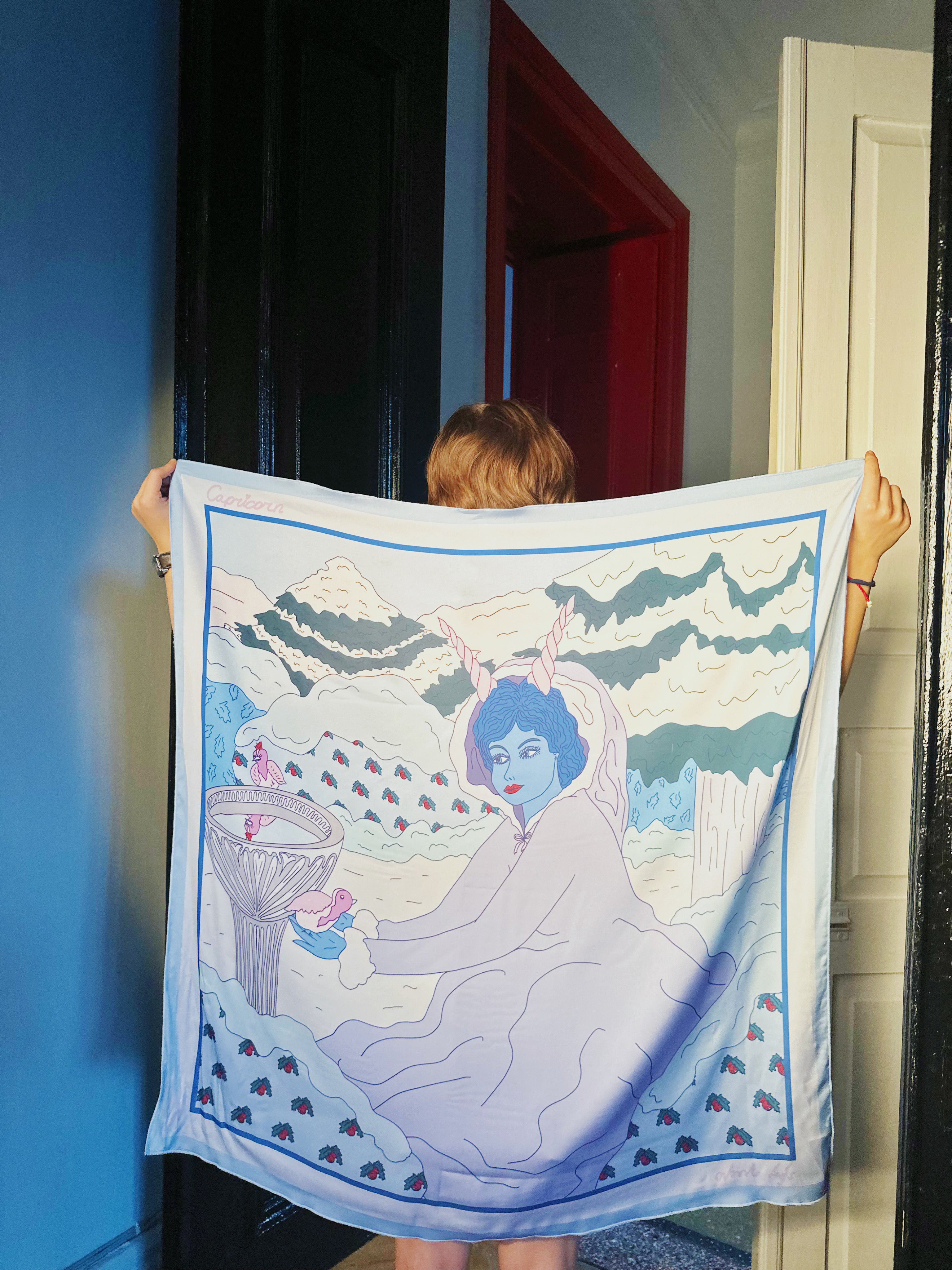 Capricorn zodiac art silk scarf hanging flat, showing detailed pastel blue and neutral-toned artwork