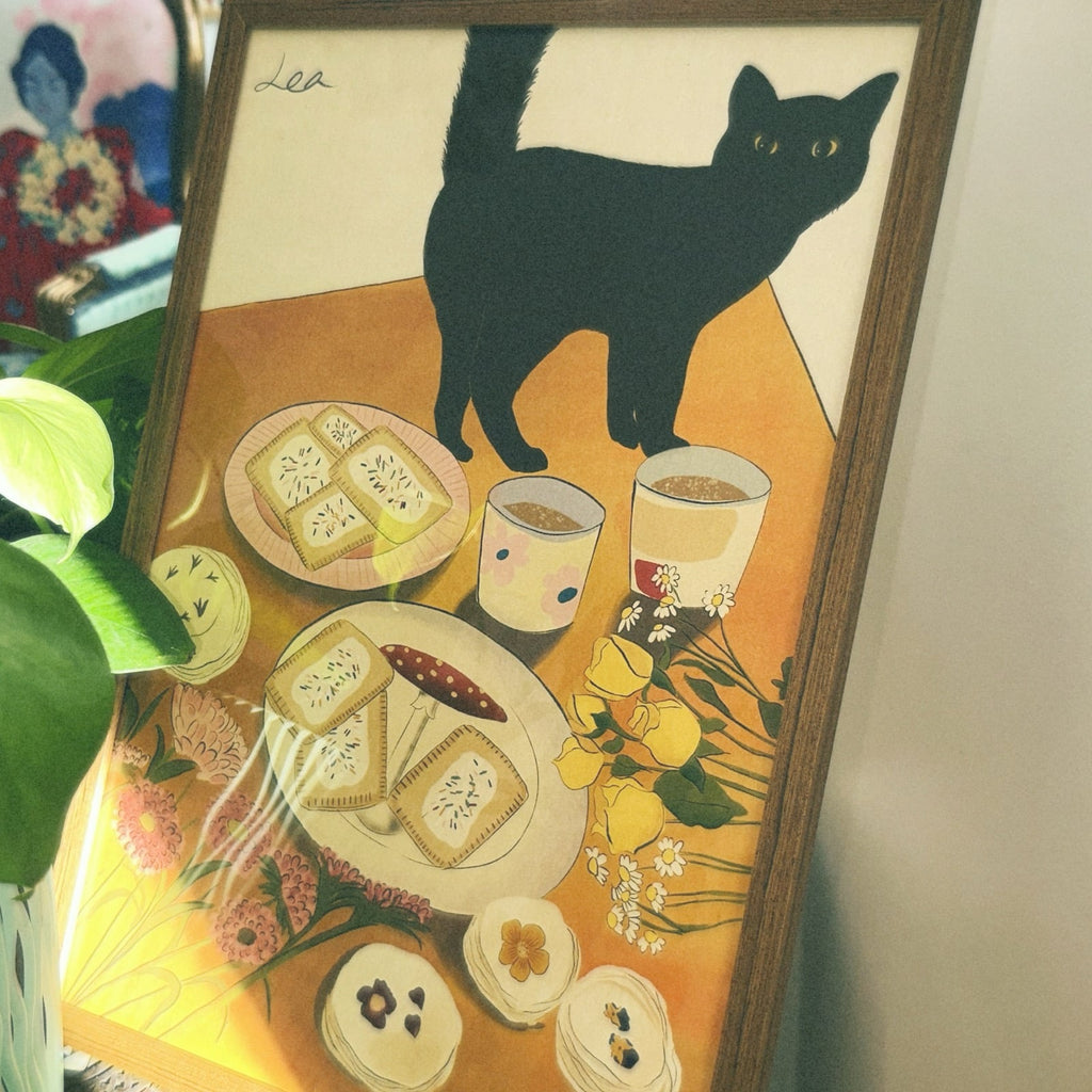 A poster featuring a black cat with a design by Lea Woo. The illustration includes plates of food, cups of tea, and floral elements in soft colors.