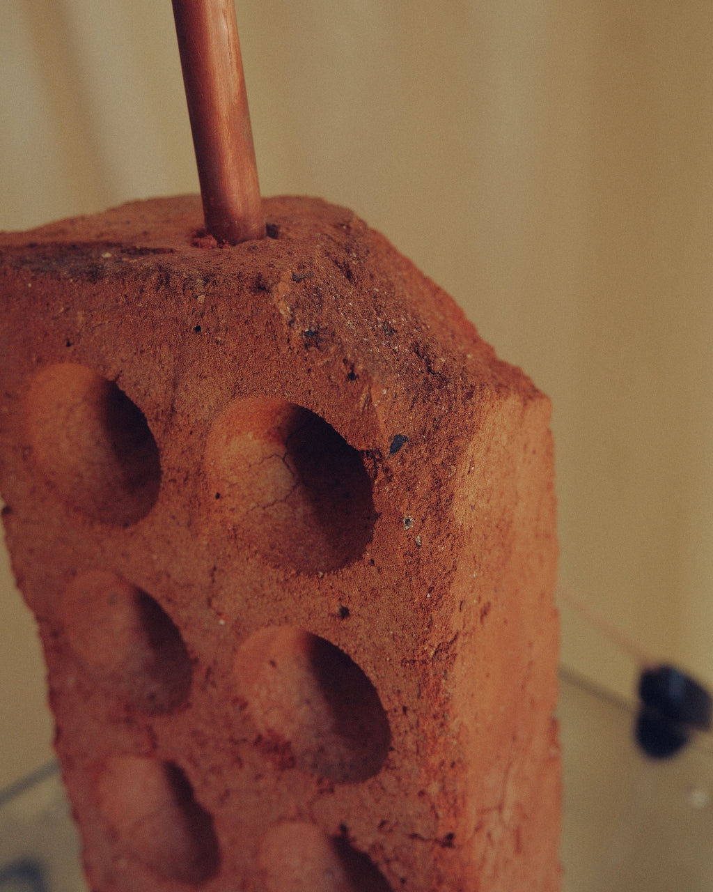 A close-up of a terracotta clay brick with circular indentations, featuring a copper-colored rod or stick inserted vertically near the top, set against a warm beige background.