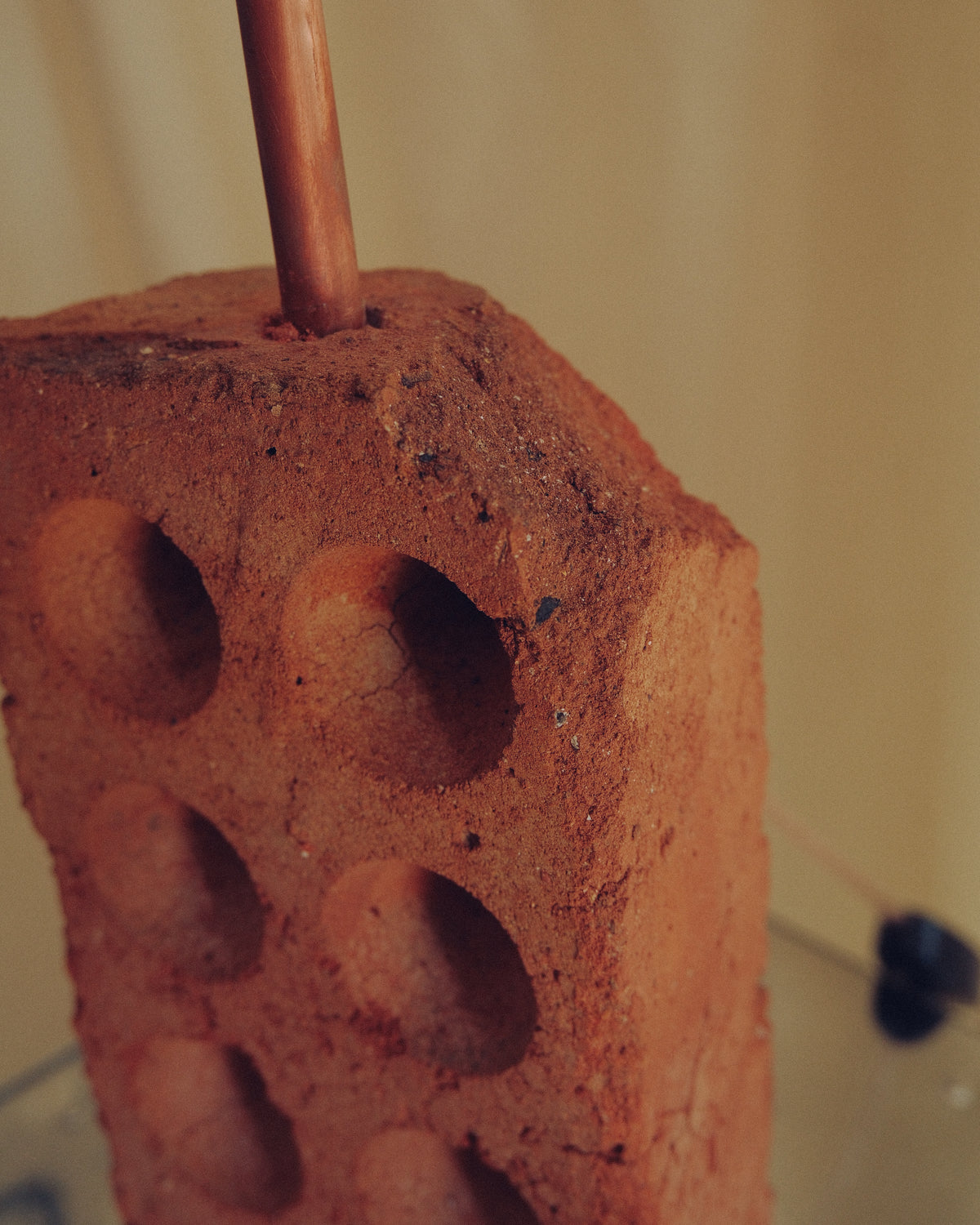 A close-up of a terracotta clay brick with circular indentations, featuring a copper-colored rod or stick inserted vertically near the top, set against a warm beige background.
