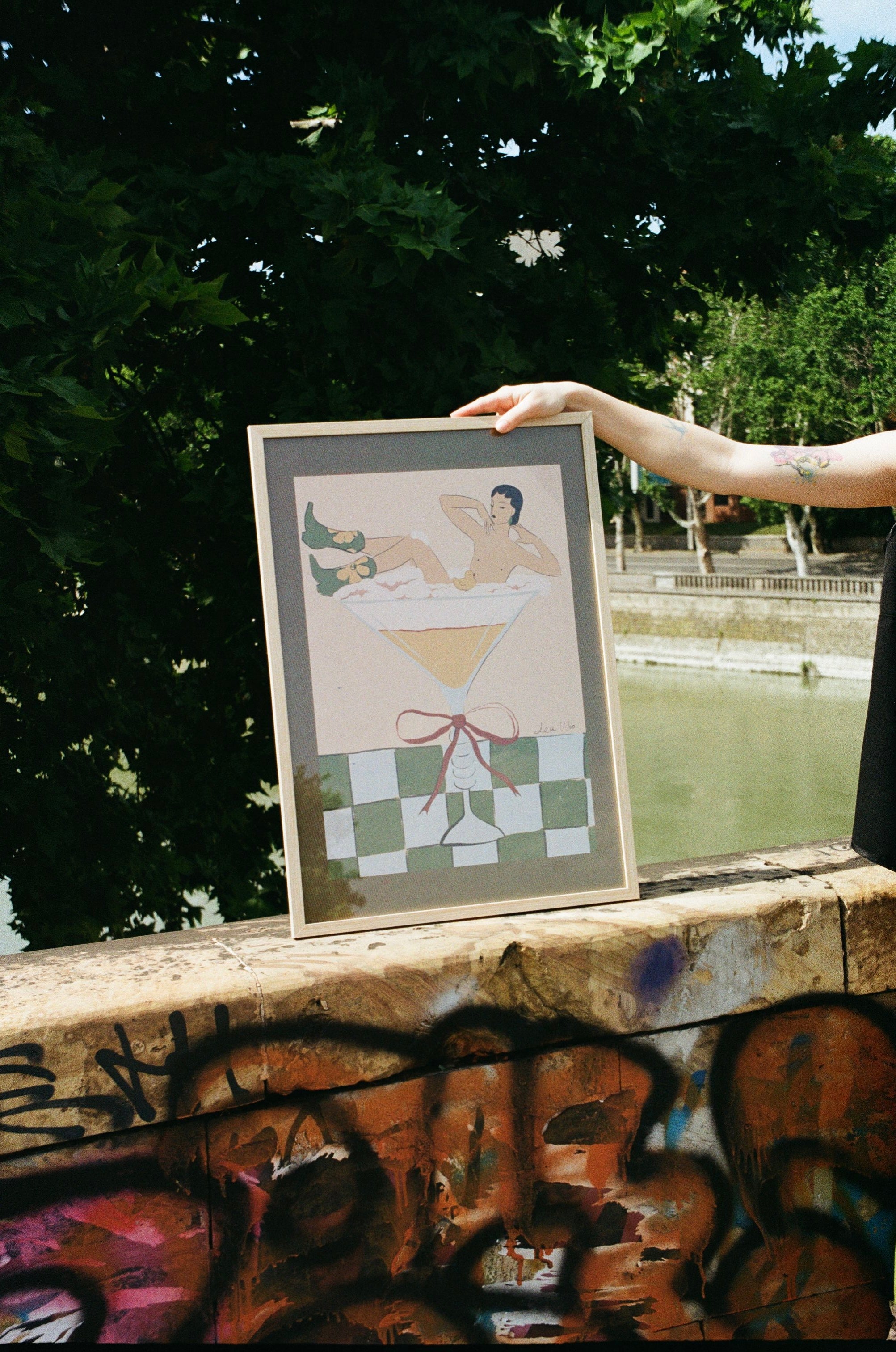 Framed artwork of a person in a bathtub held by a person against a trees in the background.