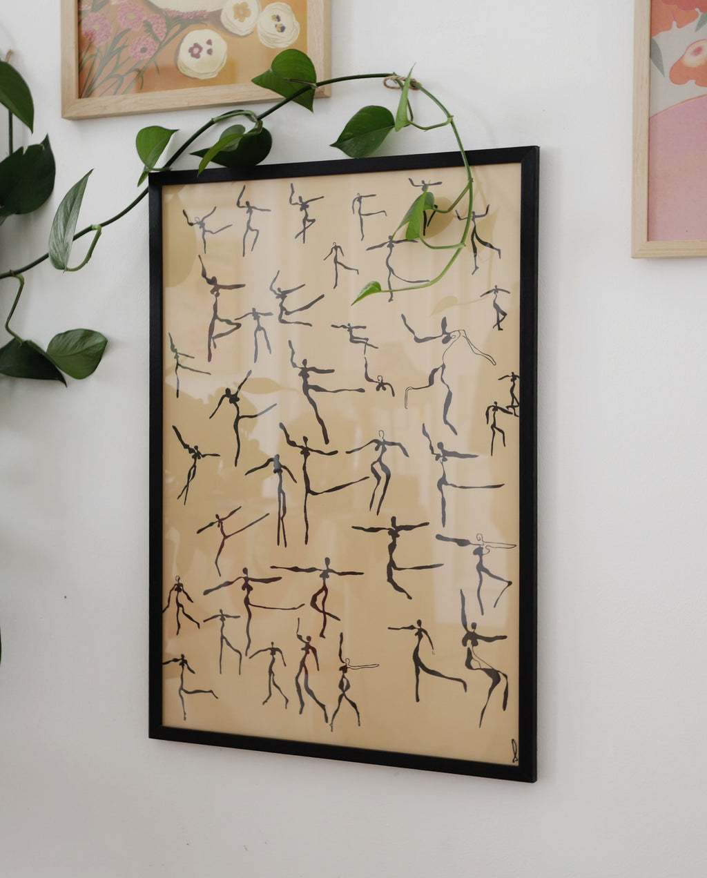 Framed artwork with stick figure patterns on a beige background, hanging on a wall with green leaves.