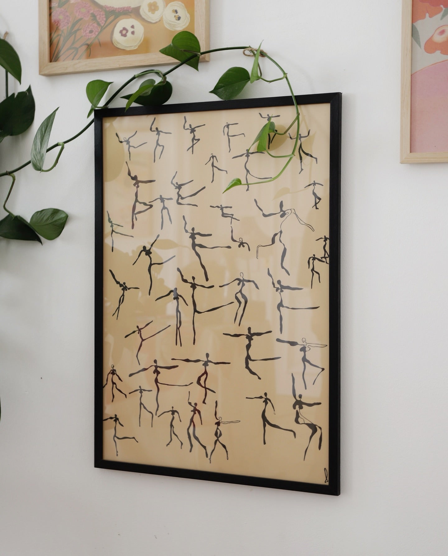 Framed artwork with stick figure patterns on a beige background, hanging on a wall with green leaves.