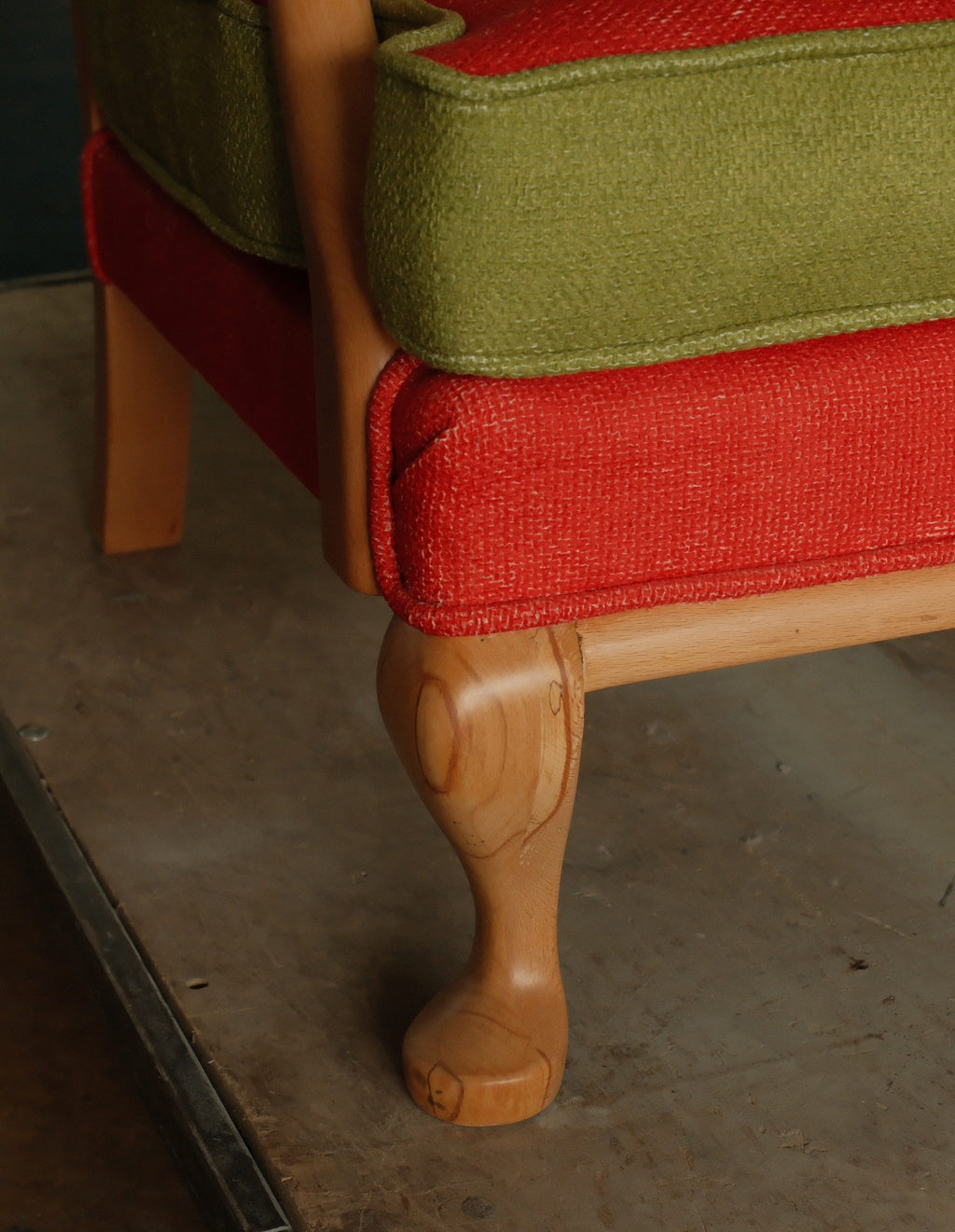 Close-up of a wooden armchair with textured red and olive green upholstery and a carved light wood leg on a concrete floor.
