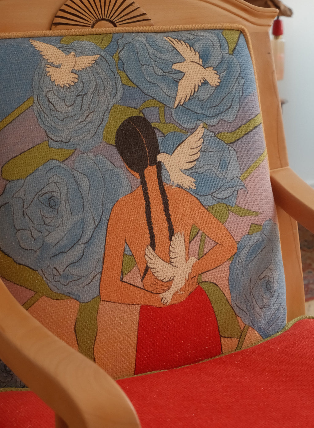 A close-up of a wooden armchair upholstered in a detailed woven tapestry featuring a woman seen from behind with braided hair, white doves, and large blue roses, with a red upholstered seat visible below.