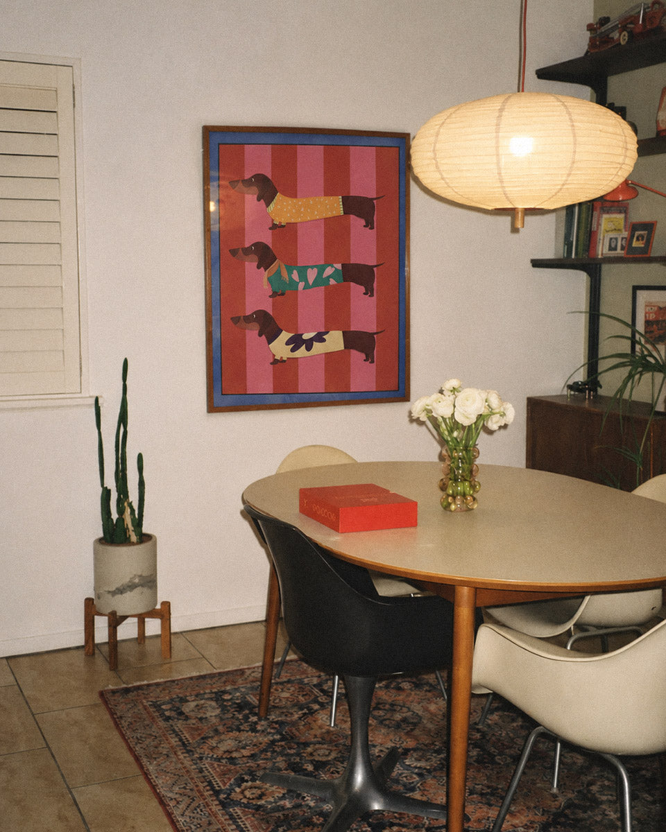 A framed art print featuring three stylized dachshund figures on pink and red striped backgrounds, hanging on a white wall in a dining room above an oval table with chairs.