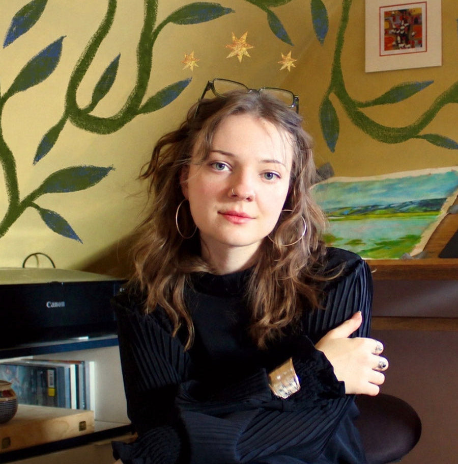 A young woman with long wavy light brown hair sits indoors wearing a black ribbed long-sleeve top and dark skirt. She is posed in front of a yellow wall decorated with green vine and leaf patterns, with a painting and shelves visible in the background.
