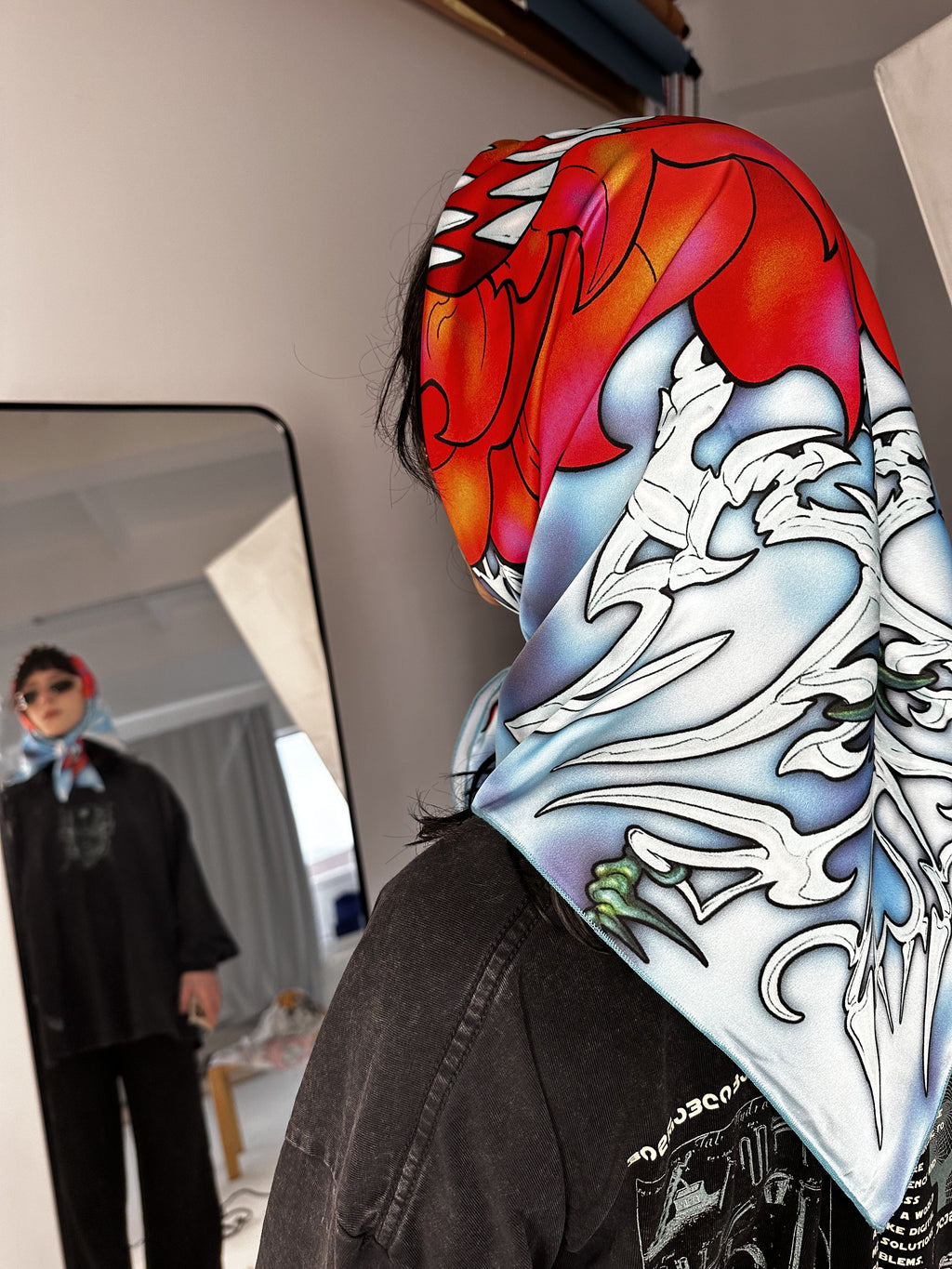 A person wearing a scarf with a colorful floral pattern, viewed from the back, with a mirror reflection showing a child in a different pose.