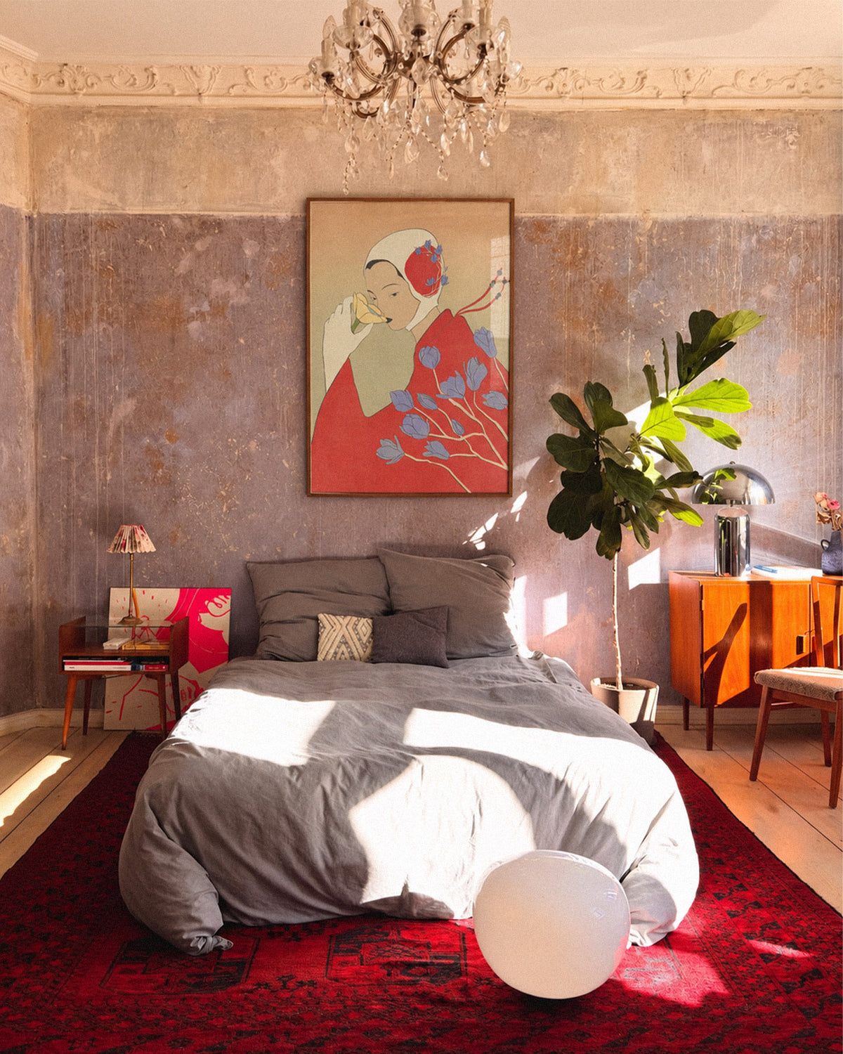 A styled bedroom interior with a bed dressed in gray bedding, a large red patterned rug, a potted green plant, an orange sideboard, and a framed abstract poster on a textured wall.