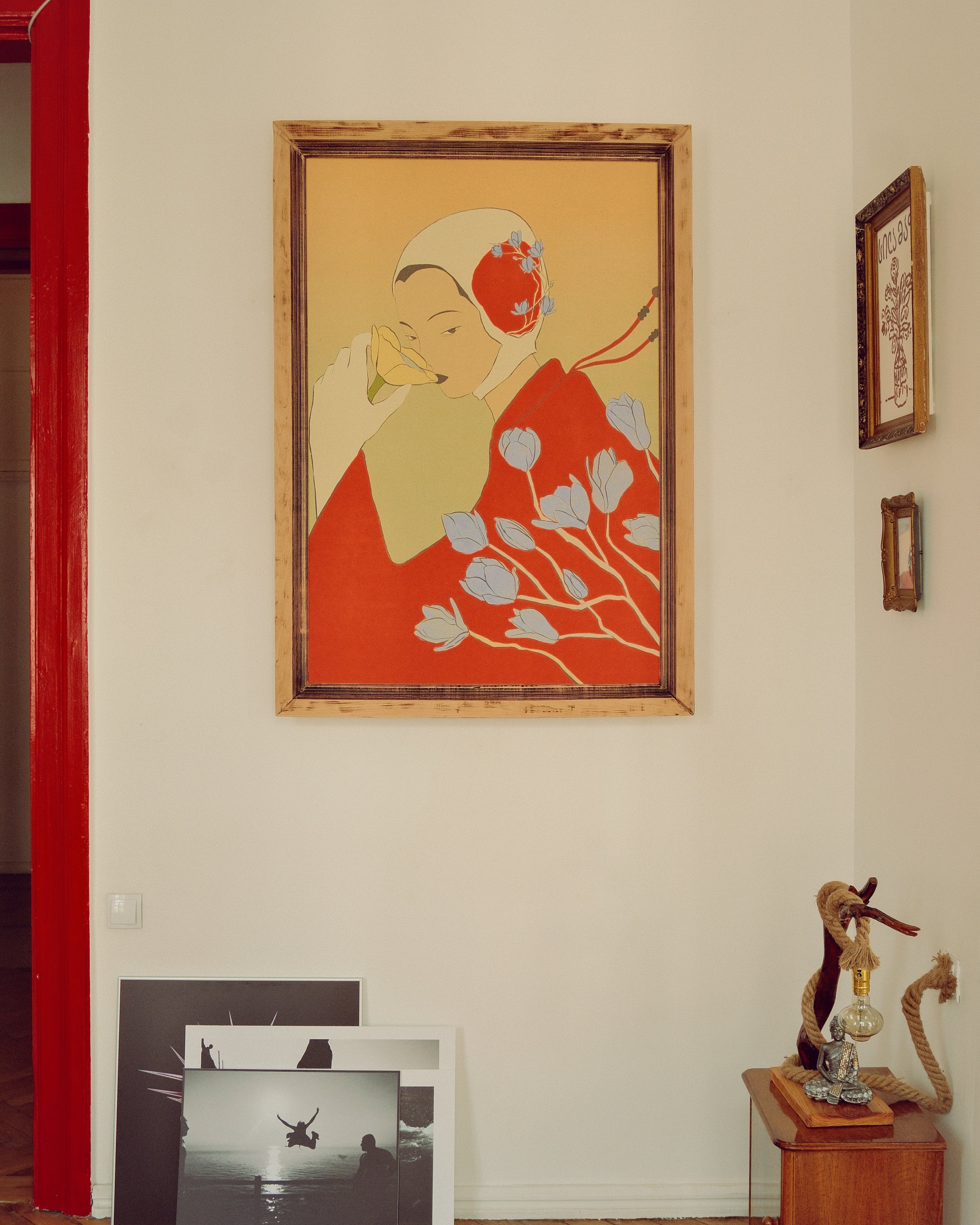 A framed art print depicting a stylized woman in a red kimono with white flowers, hanging on a cream-colored wall in a room with other framed pictures and a wooden side table.