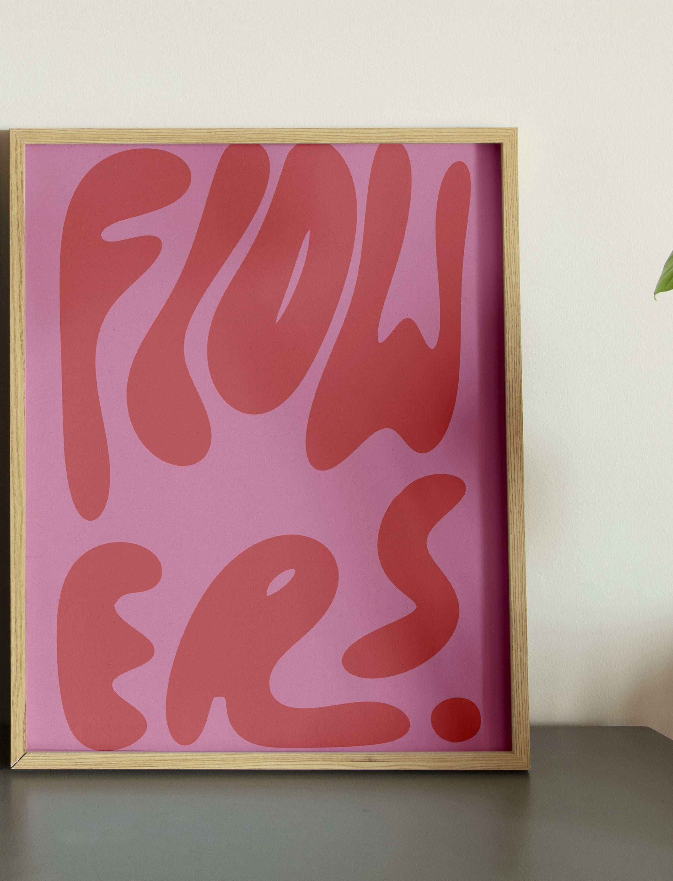 A framed poster with the word 'FLOWERS' in stylized lettering, predominantly in one color with a pink background.