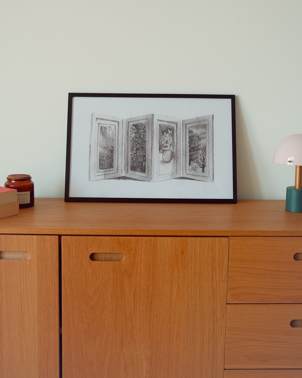 A framed black and white sketch of four open book pages or doors, displayed on a light wood sideboard against a pale wall.