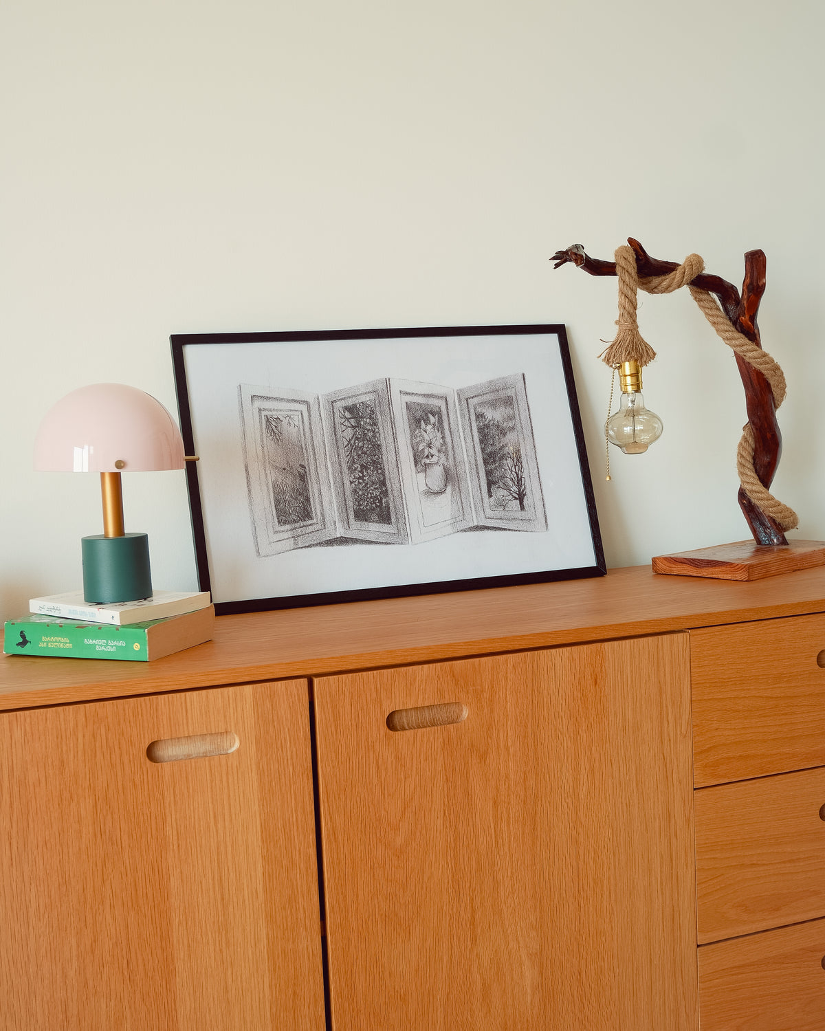 A framed black and white art print depicting open book pages, styled on a light wood dresser alongside a pink dome lamp and a rope-wrapped branch light against a cream wall.