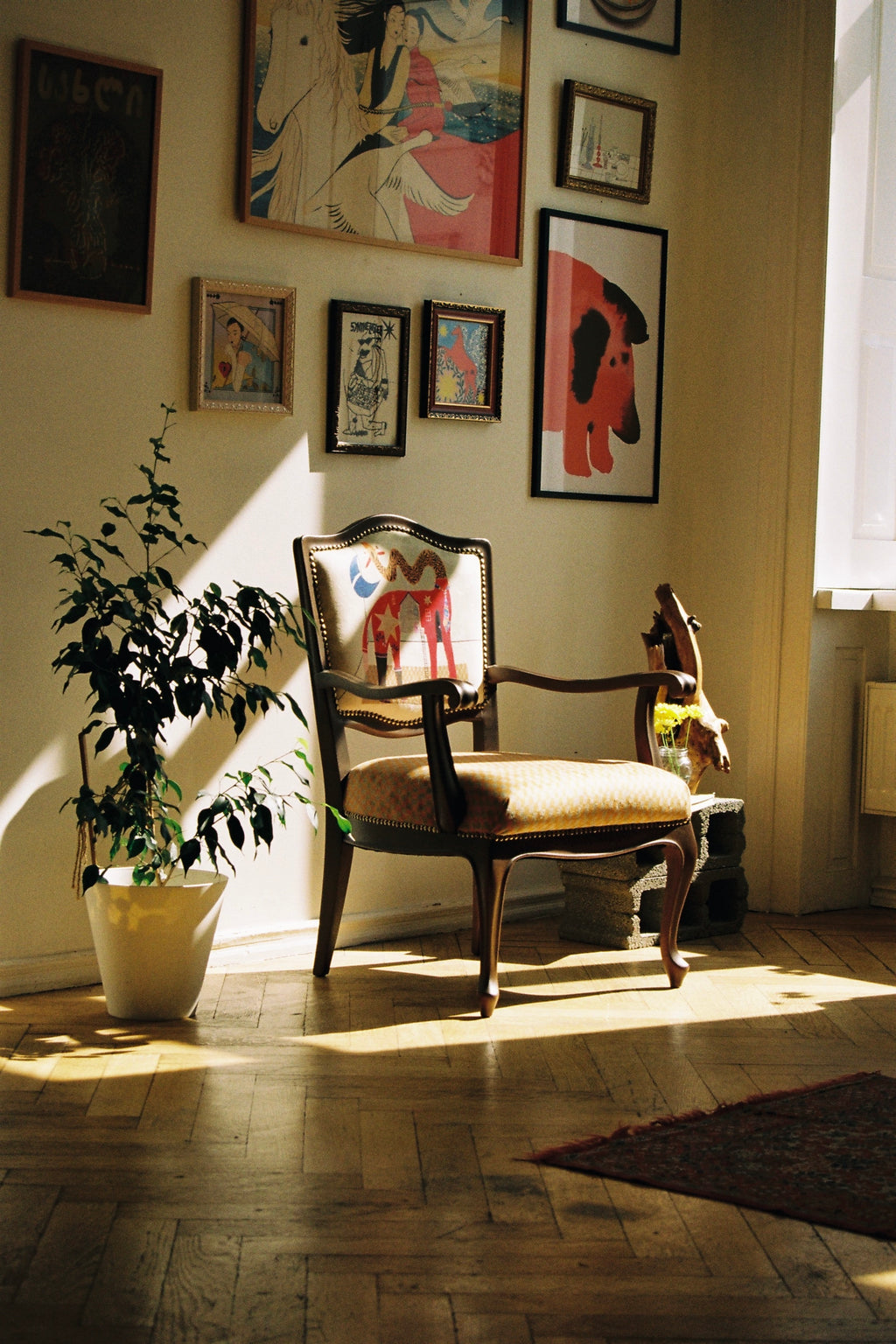 A vintage wooden armchair with a beige upholstered seat and back, styled in a sunlit room with framed art on the wall, a potted plant, and warm natural light streaming across the floor.