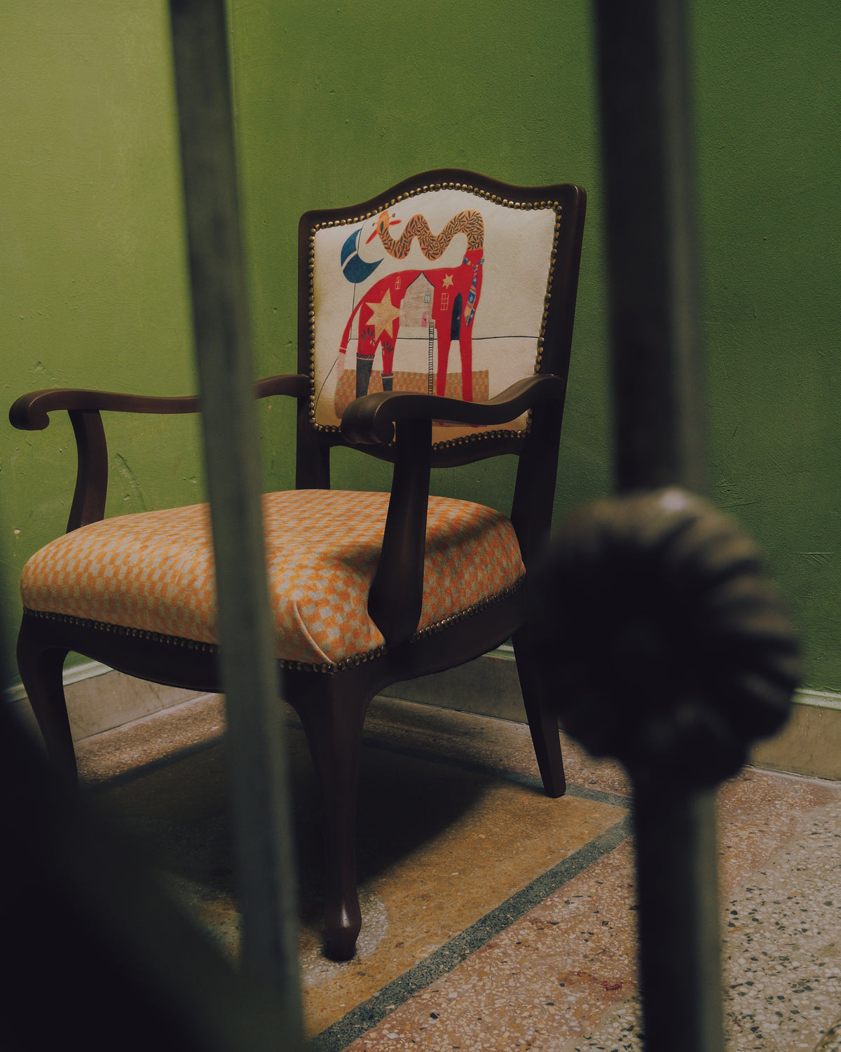 An antique-style wooden armchair with a cream upholstered backrest featuring a colorful abstract painting and a patterned orange-and-white cushioned seat, set against a green wall.
