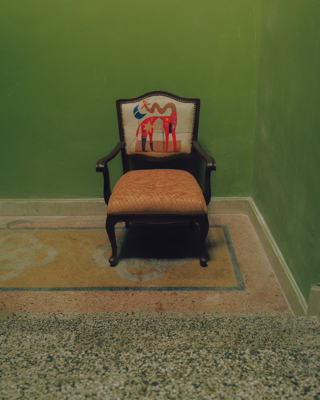 A vintage-style upholstered armchair with a dark wood frame, patterned orange-brown seat cushion, and a cream backrest cushion featuring a colorful abstract horse design, set in a green room corner on a patterned rug.