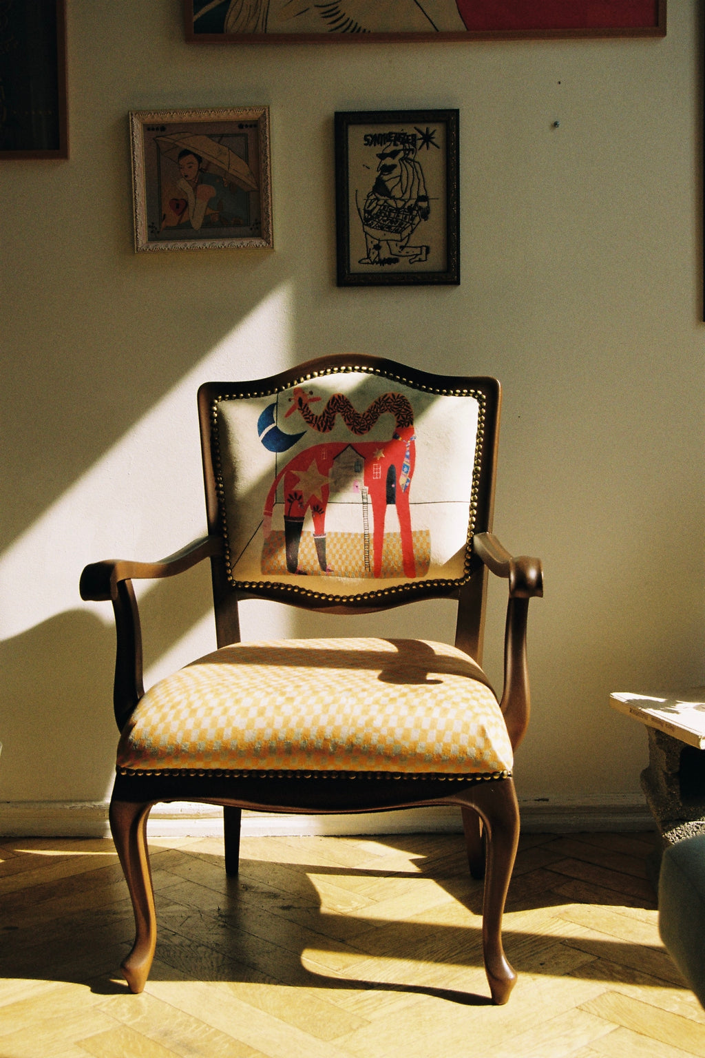 A vintage-style wooden armchair with a patterned yellow and white checkered seat cushion and a colorful horse-back motif on the backrest, placed in a sunlit room on a parquet floor.