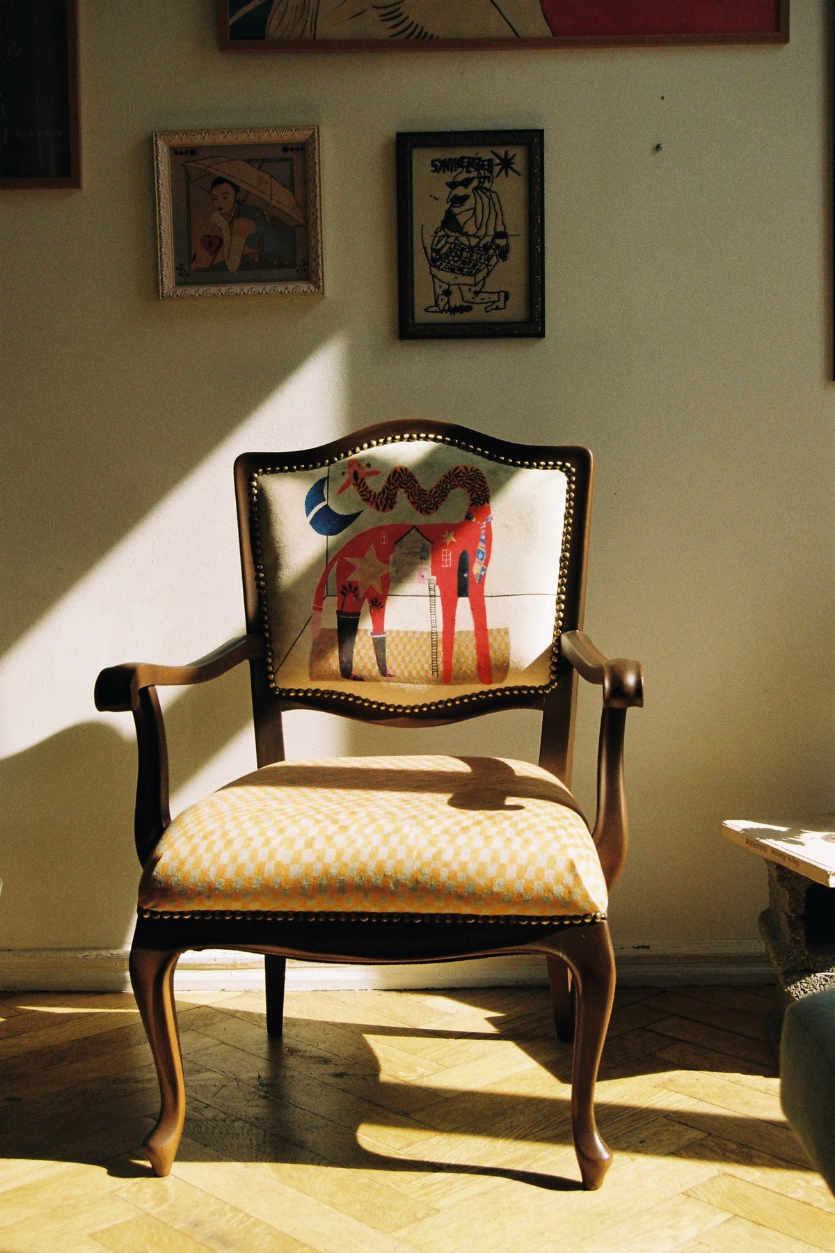 A vintage-style wooden armchair with a patterned yellow and white checkered seat cushion and a colorful horse-back motif on the backrest, placed in a sunlit room on a parquet floor.