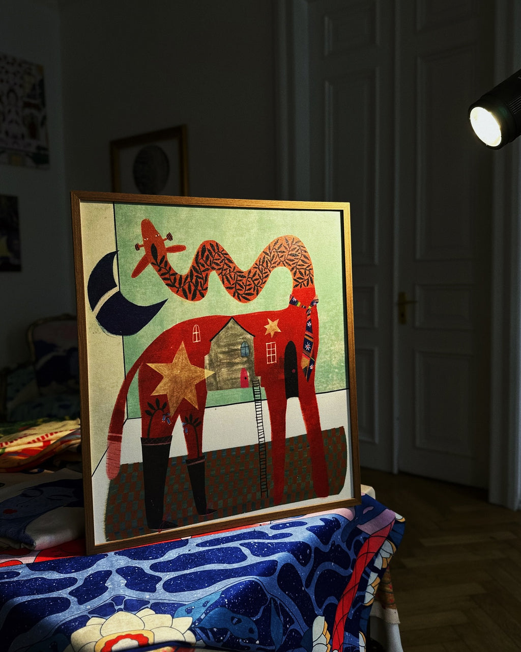 Framed giraffe art poster in warm, colorful style, displayed on a table in an interior space. Contemporary print by DAE artist.
