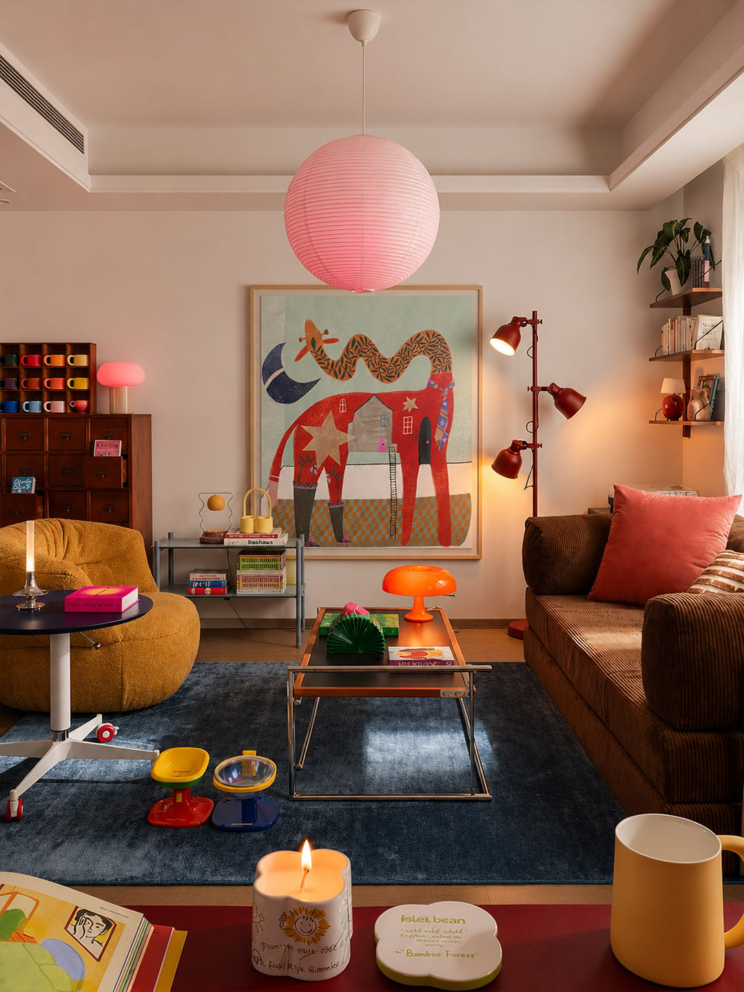 A cozy, colorful living room featuring a brown sofa with a pink pillow, a mustard yellow armchair, a blue rug, and a large colorful poster by DAE on the wall. A pink paper lantern hangs from the ceiling, and various home decor items are styled throughout the space.