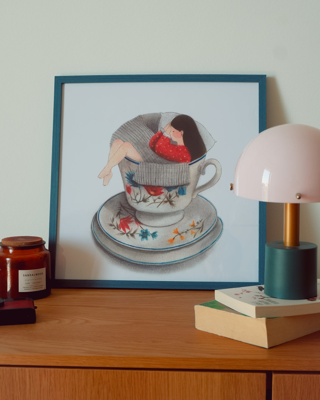 A framed art print depicting a woman curled up inside a teacup and saucer, styled on a wooden table next to a pink dome lamp and a candle.