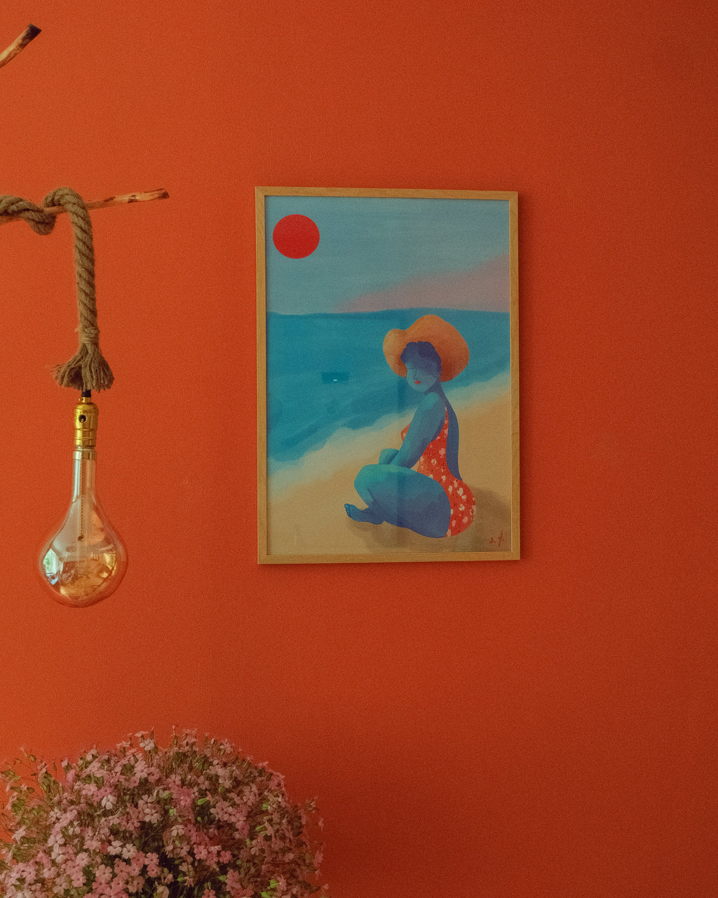 Framed artwork of a woman on a beach with a red sun, hanging on an orange wall.