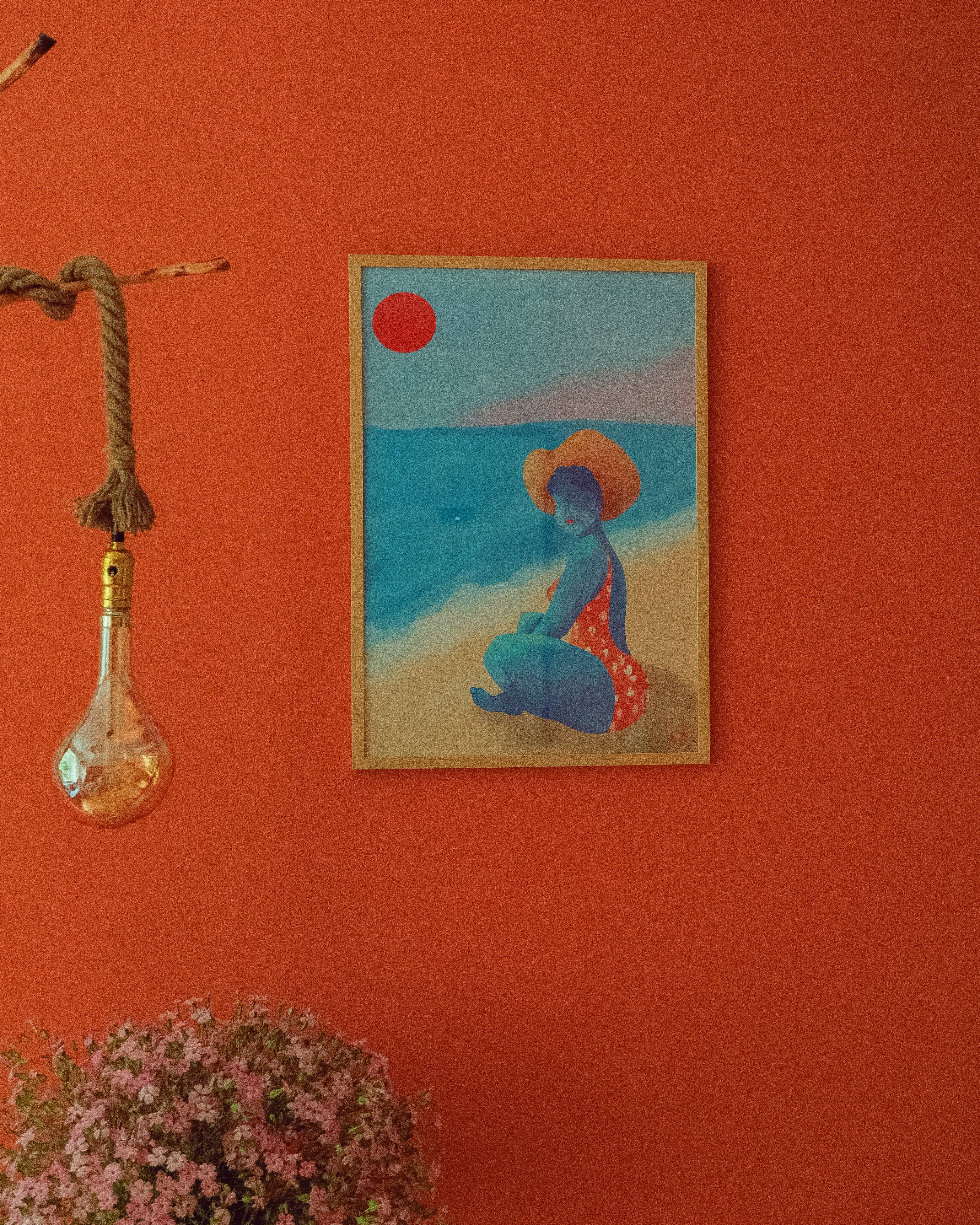 Framed artwork of a woman on a beach with a red sun, hanging on an orange wall.