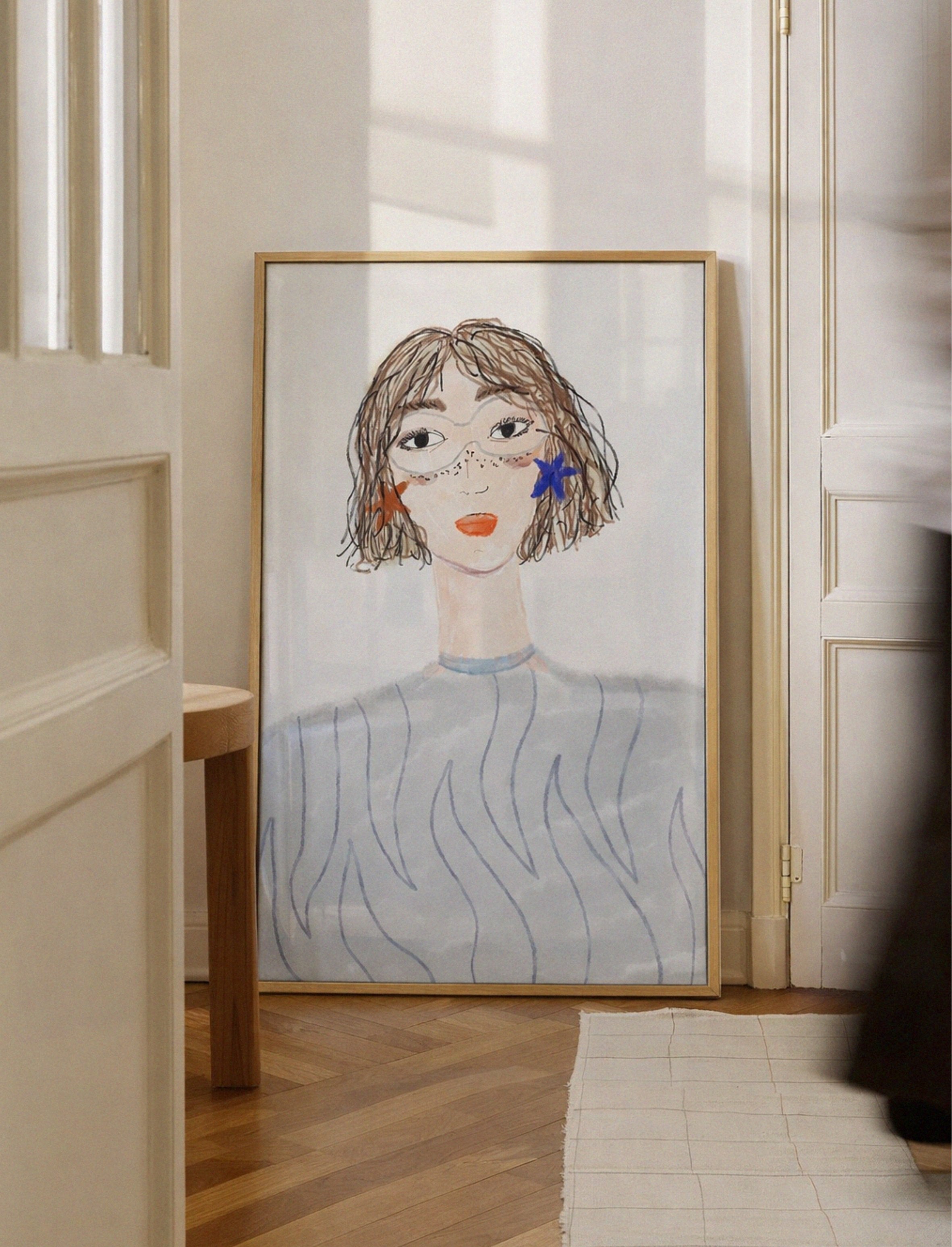 Framed artwork of a stylized portrait on a wall in a room with wooden floor and white walls.