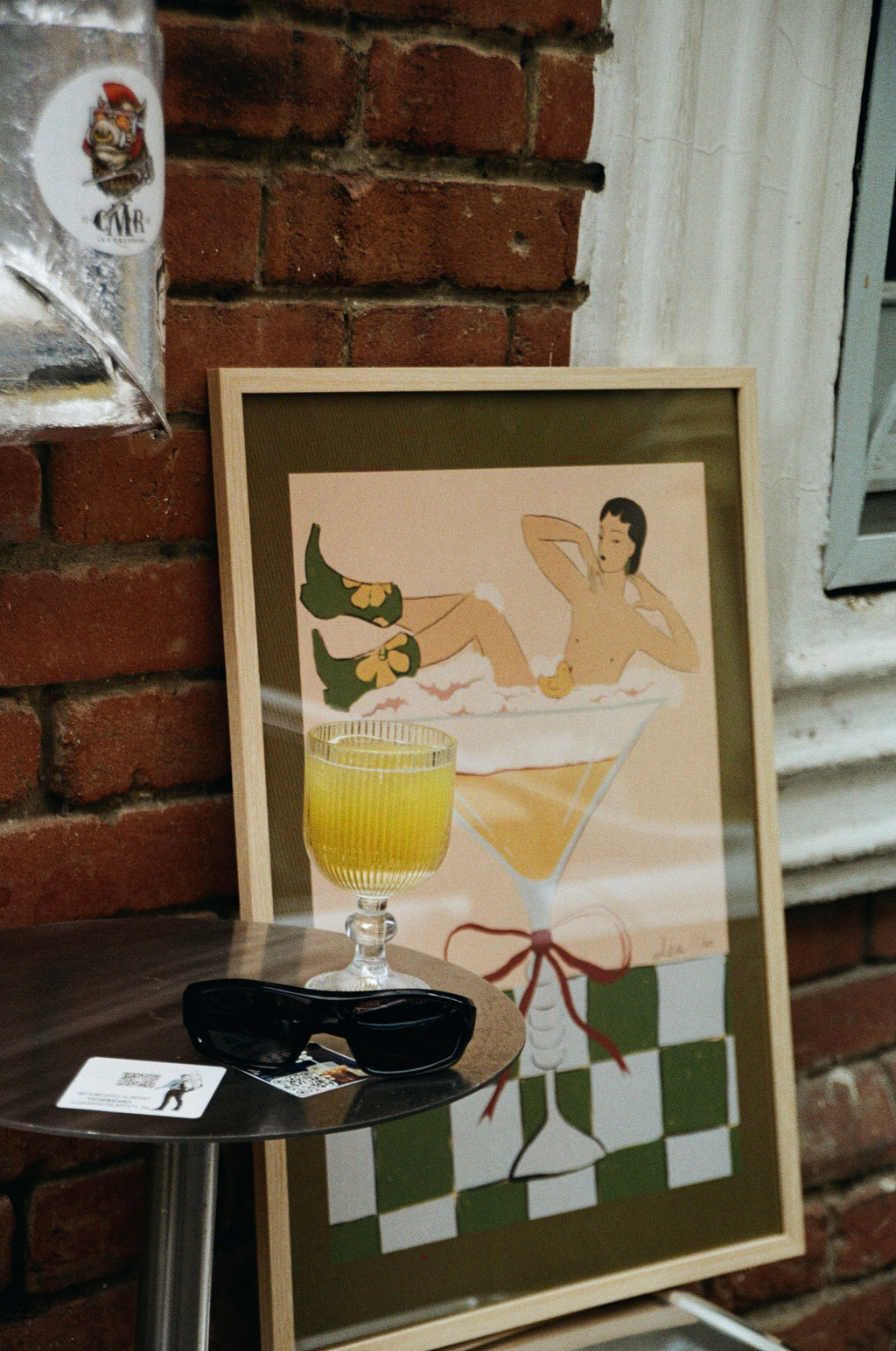 Framed illustration of a woman in warm tones placed on a table outdoors near a brick wall. Poetic, soft style artwork by Lea Woo.
