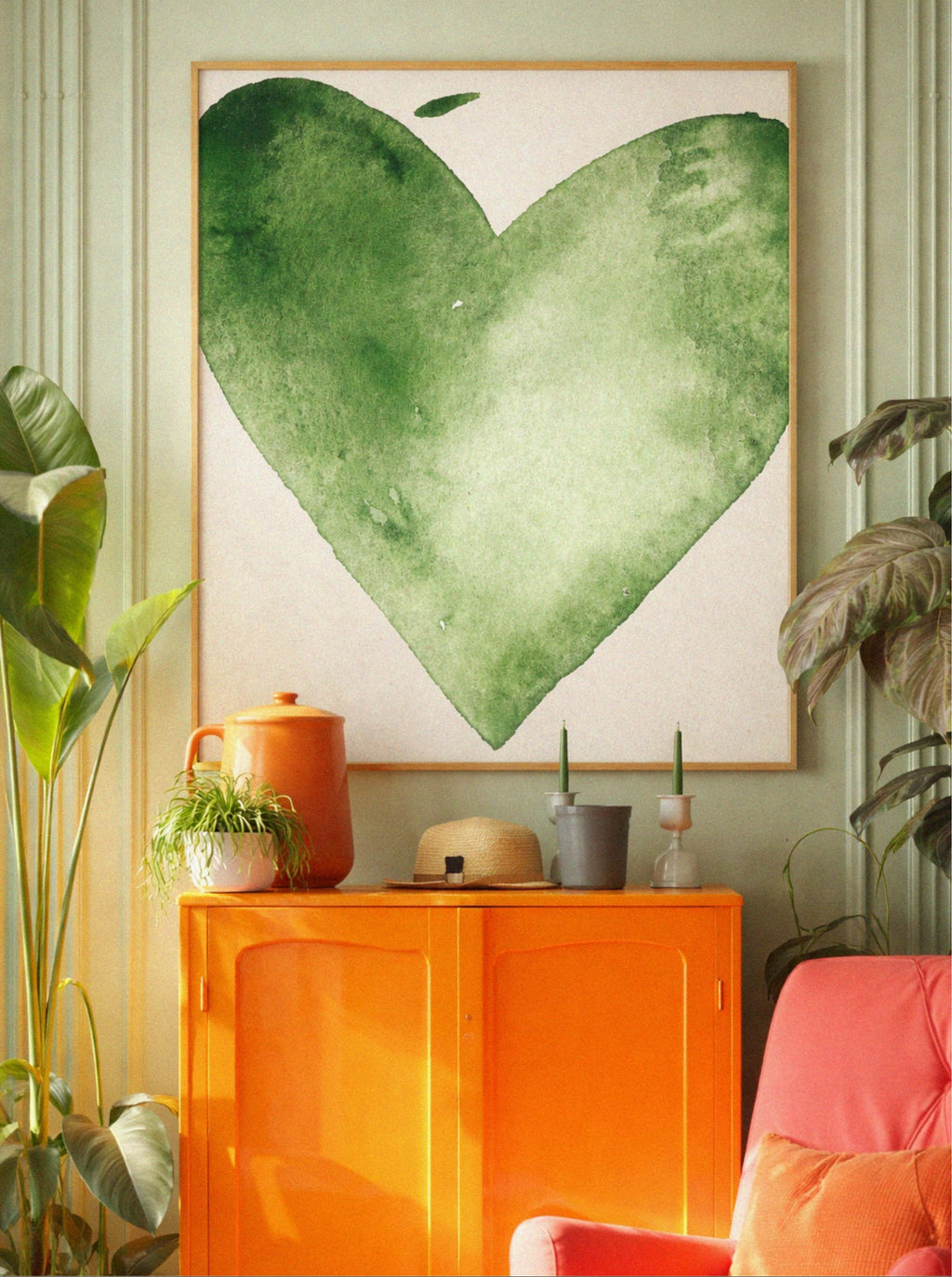 Heart-shaped green artwork on a wall with an orange cabinet and pink chair in a room.